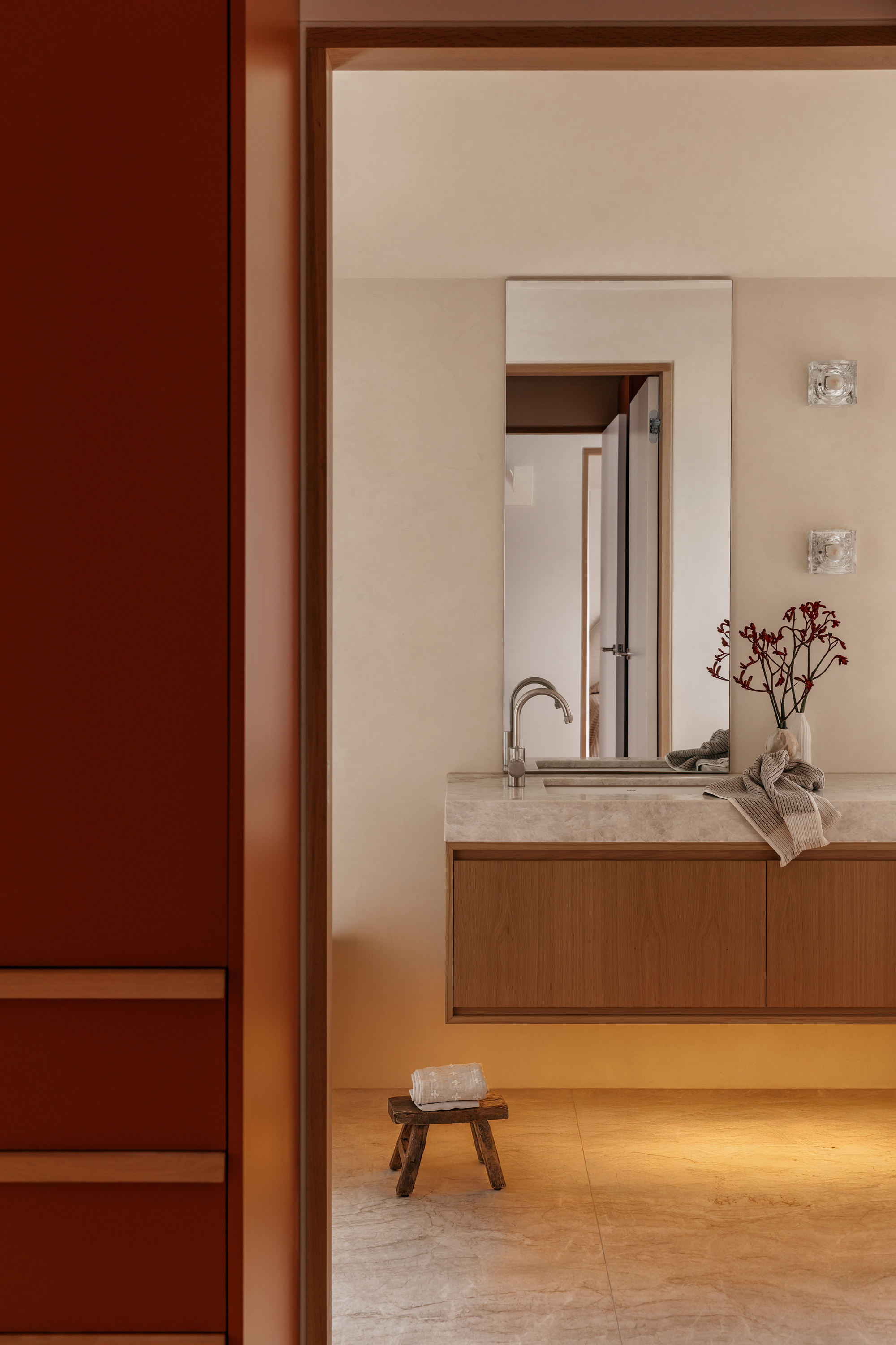 Ensuite seen through a doorway, with a floating oak vanity, stone top, tall mirror and small timber stool in warm light