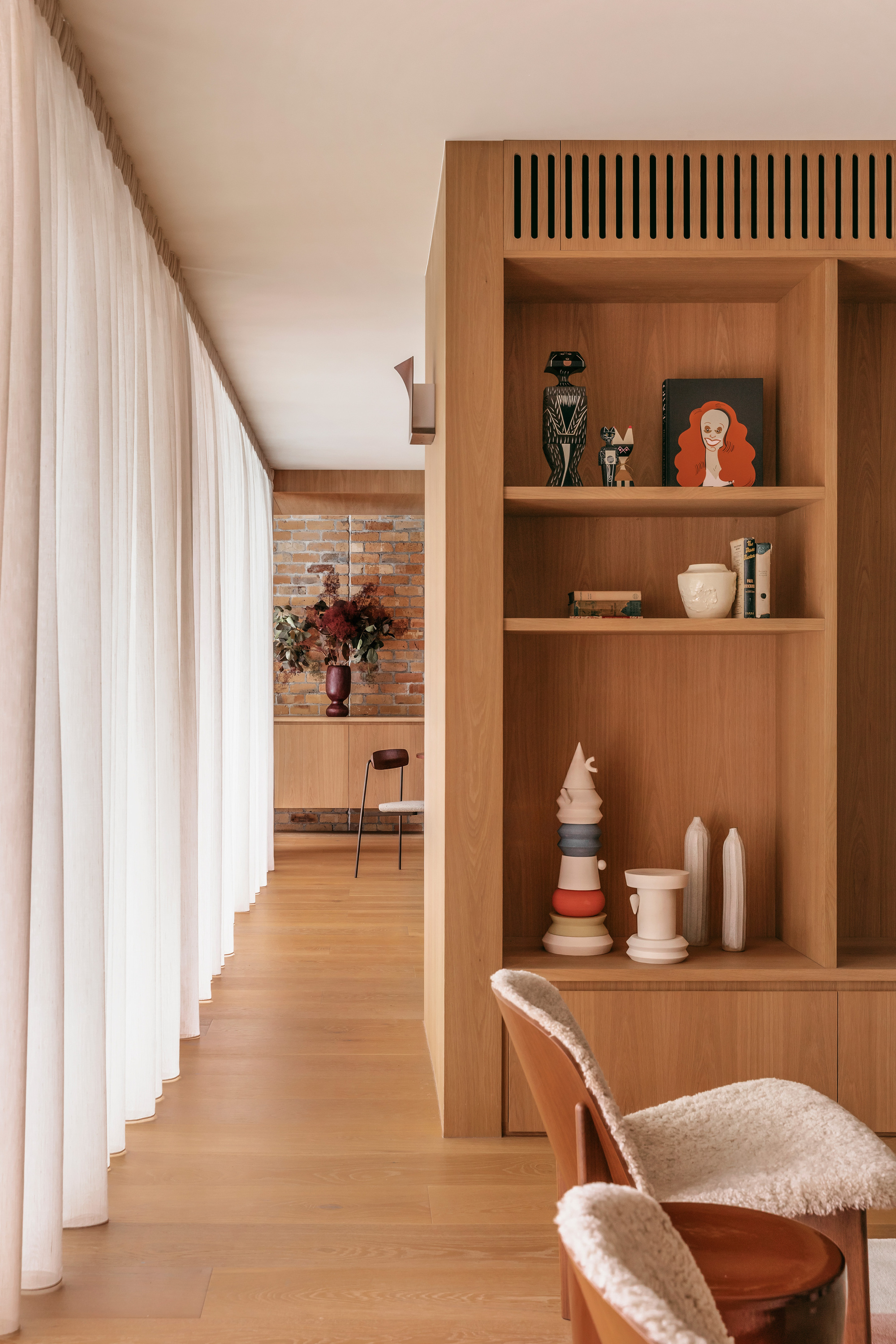 Oak shelving unit with ceramics and framed portrait beside a sheer curtain, leading toward an exposed brick wall and timber floor