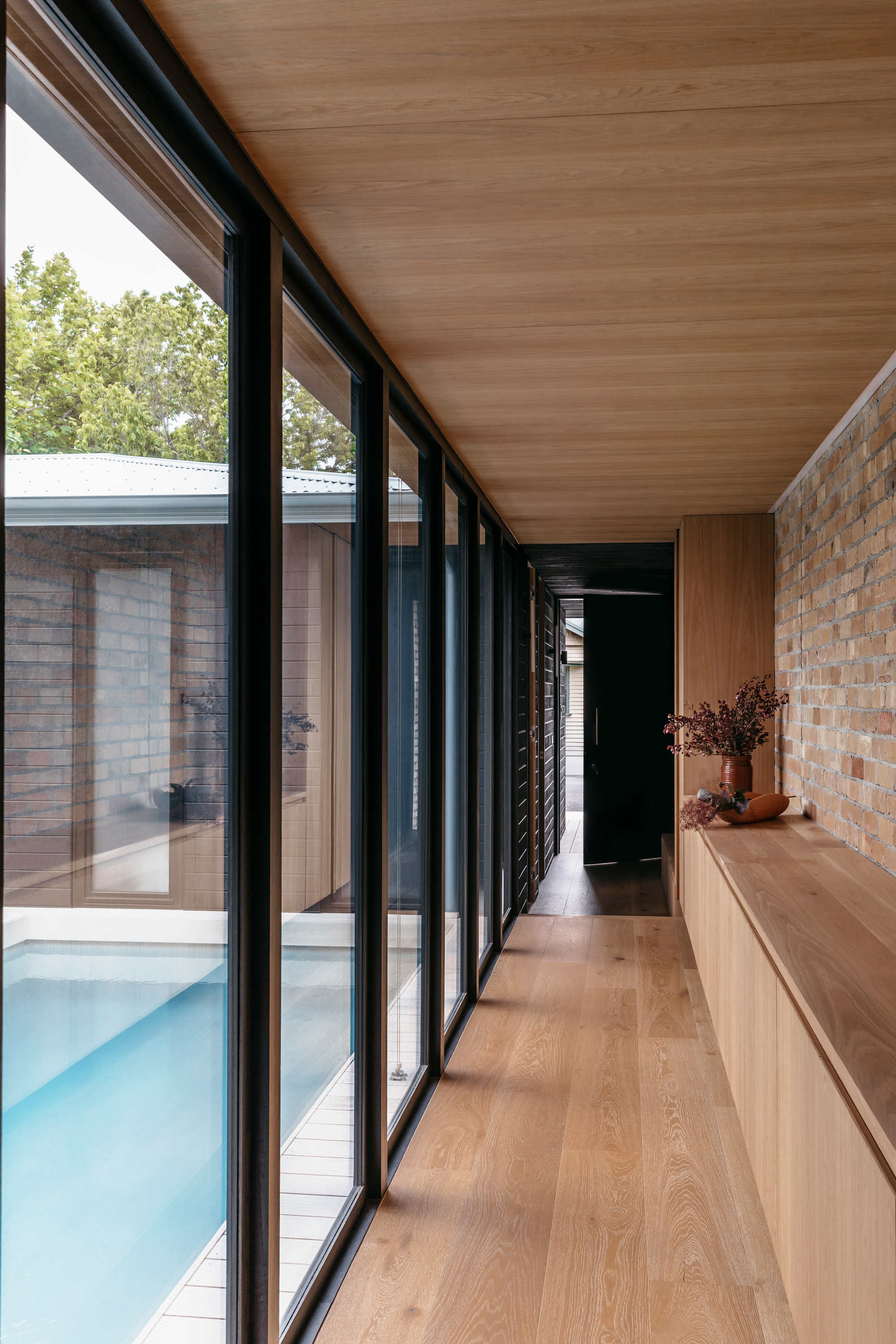 Timber-lined corridor with black-framed glazing facing a courtyard plunge pool, brick wall and built-in oak bench with dried flowers on the right