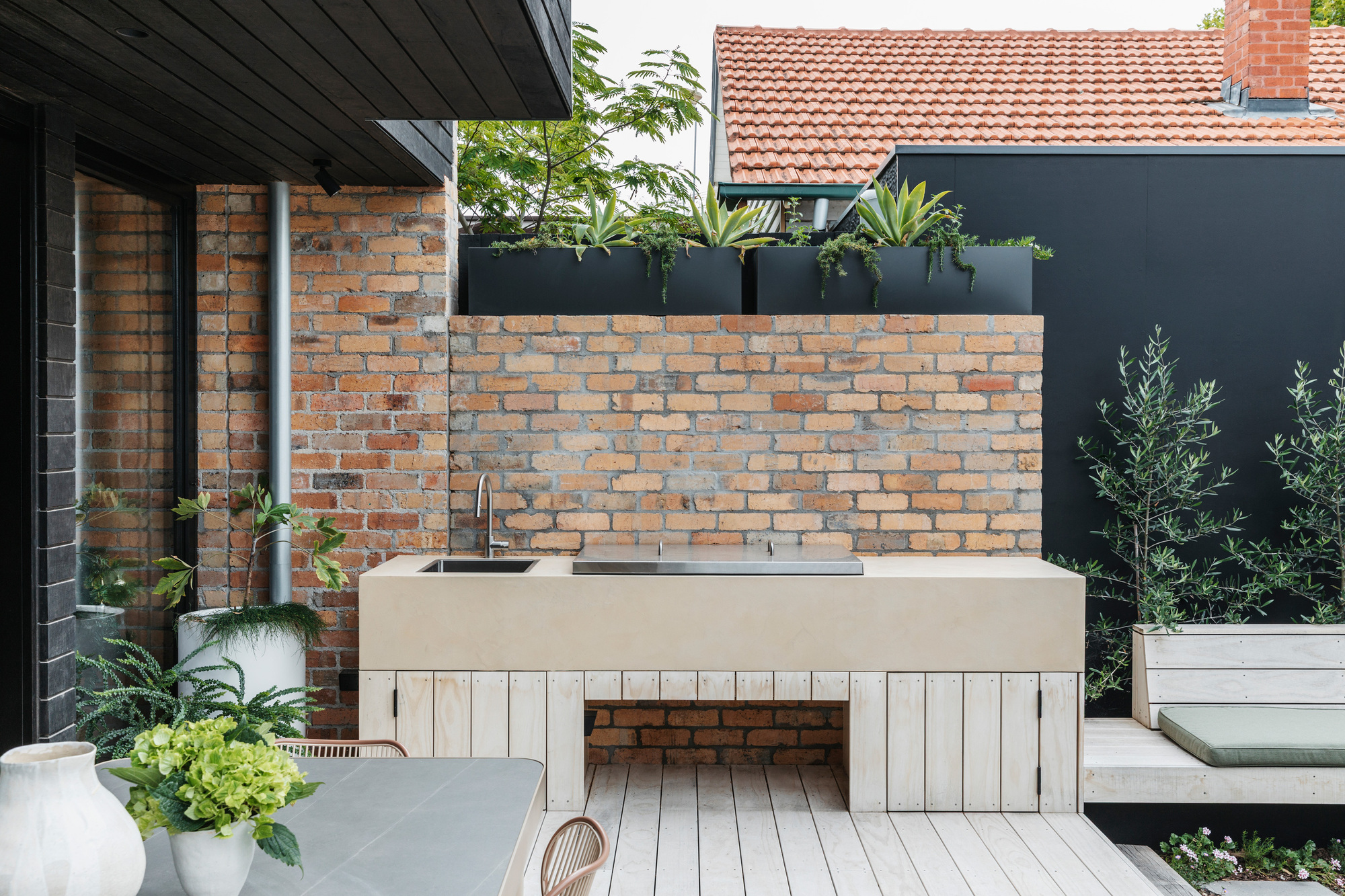 Outdoor kitchen with a pale stone benchtop and sink against a reclaimed brick wall, with planters and olive trees behind