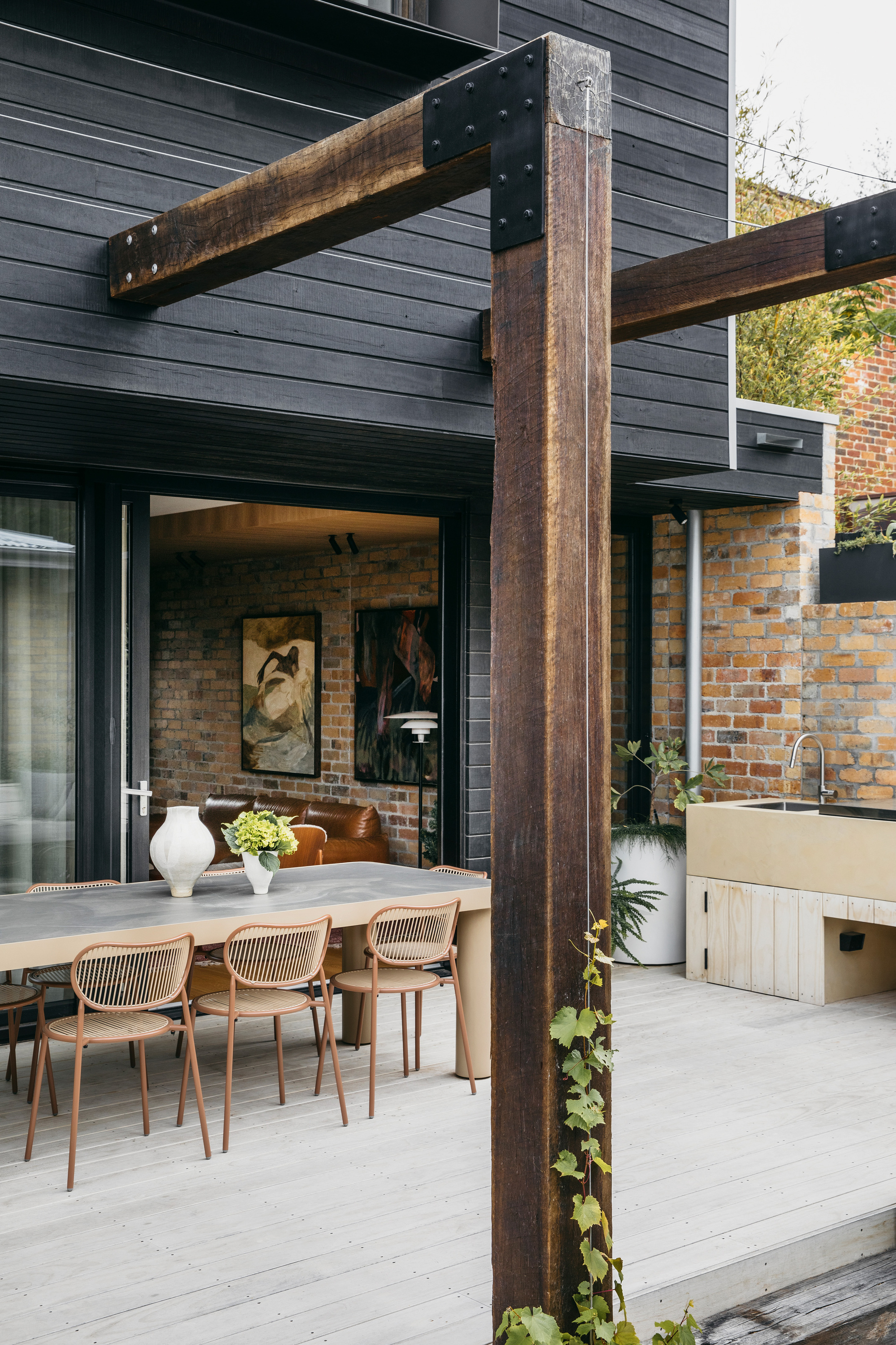 Dark timber post and steel bracket frame a deck dining table with rust-toned chairs, open glazing and a creeper climbing the post