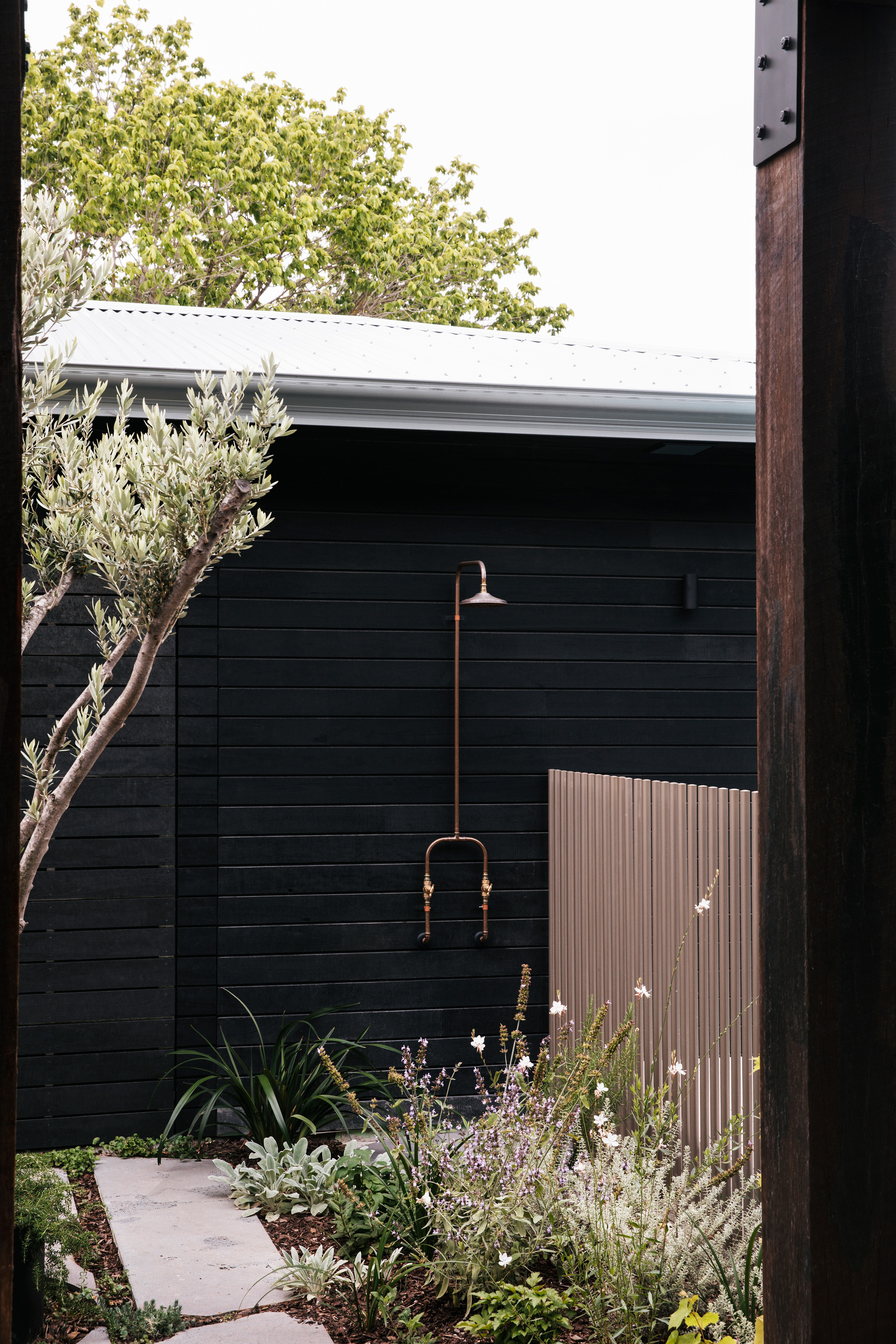 Outdoor copper shower on a black timber-clad wall, with olive branches, flowering plants and a stone path in daylight