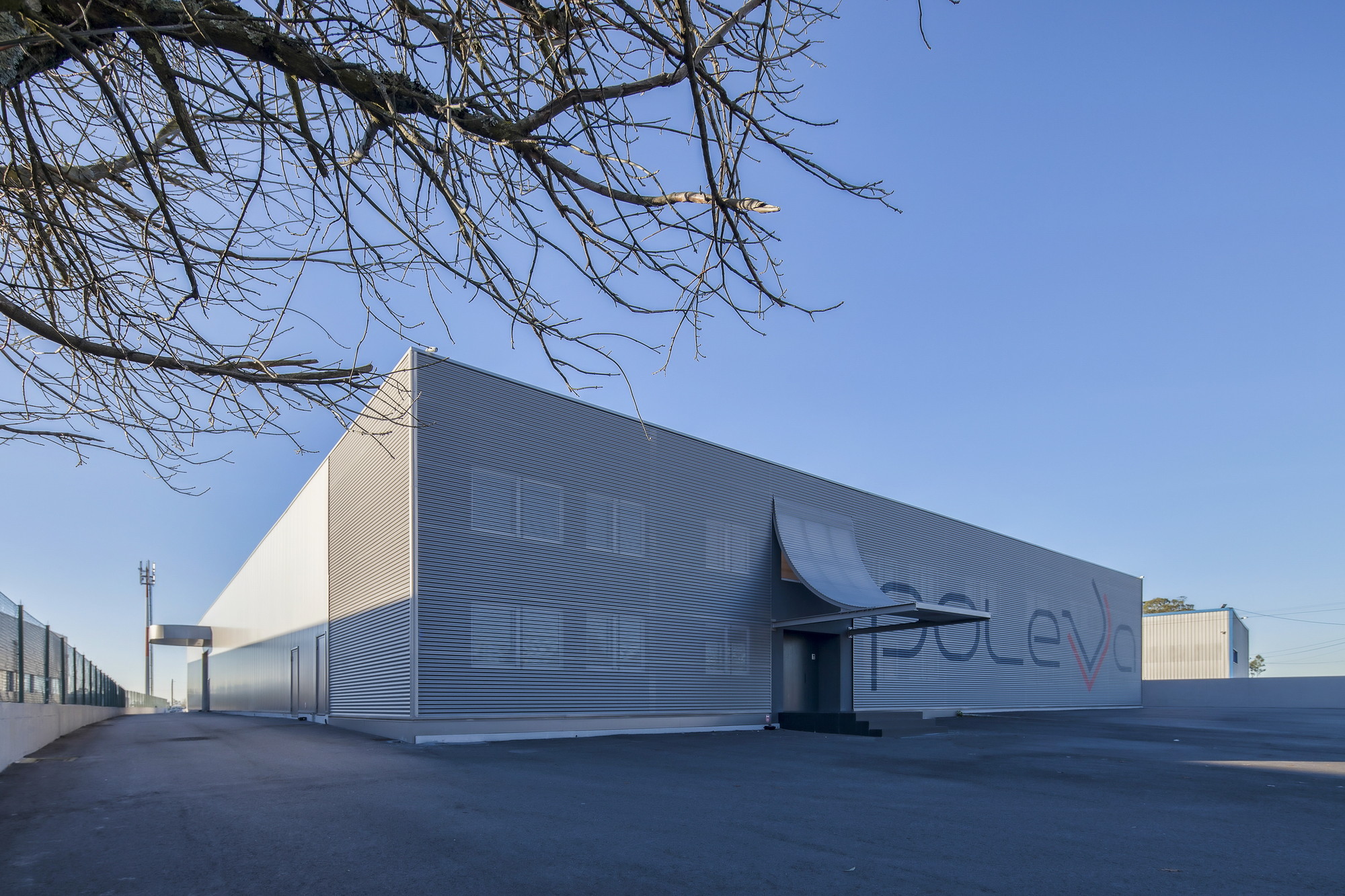 Corrugated metal warehouse facade with a curved entrance canopy and painted lettering, seen under a clear blue sky past bare branches