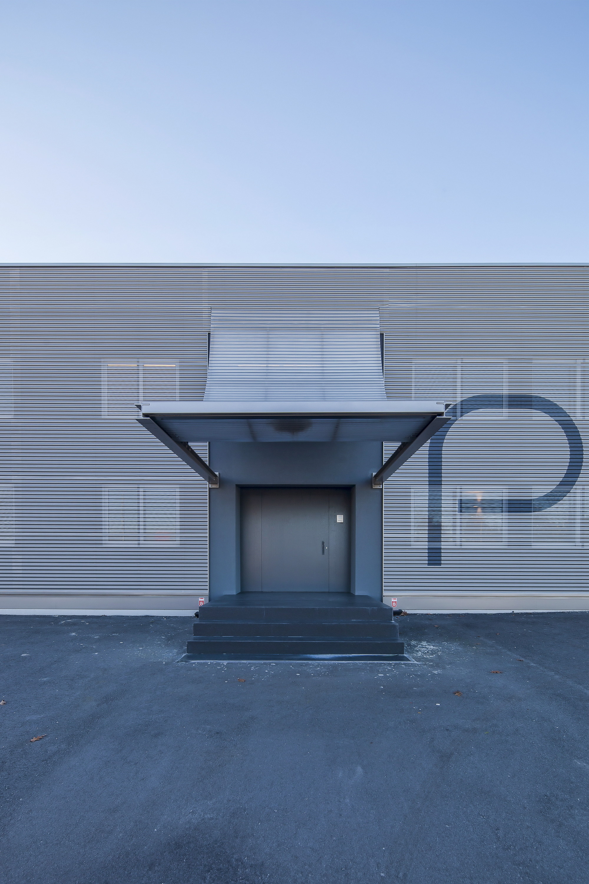 Frontal view of the entrance door beneath a curved metal canopy, with dark steps and a large painted letter on the cladding