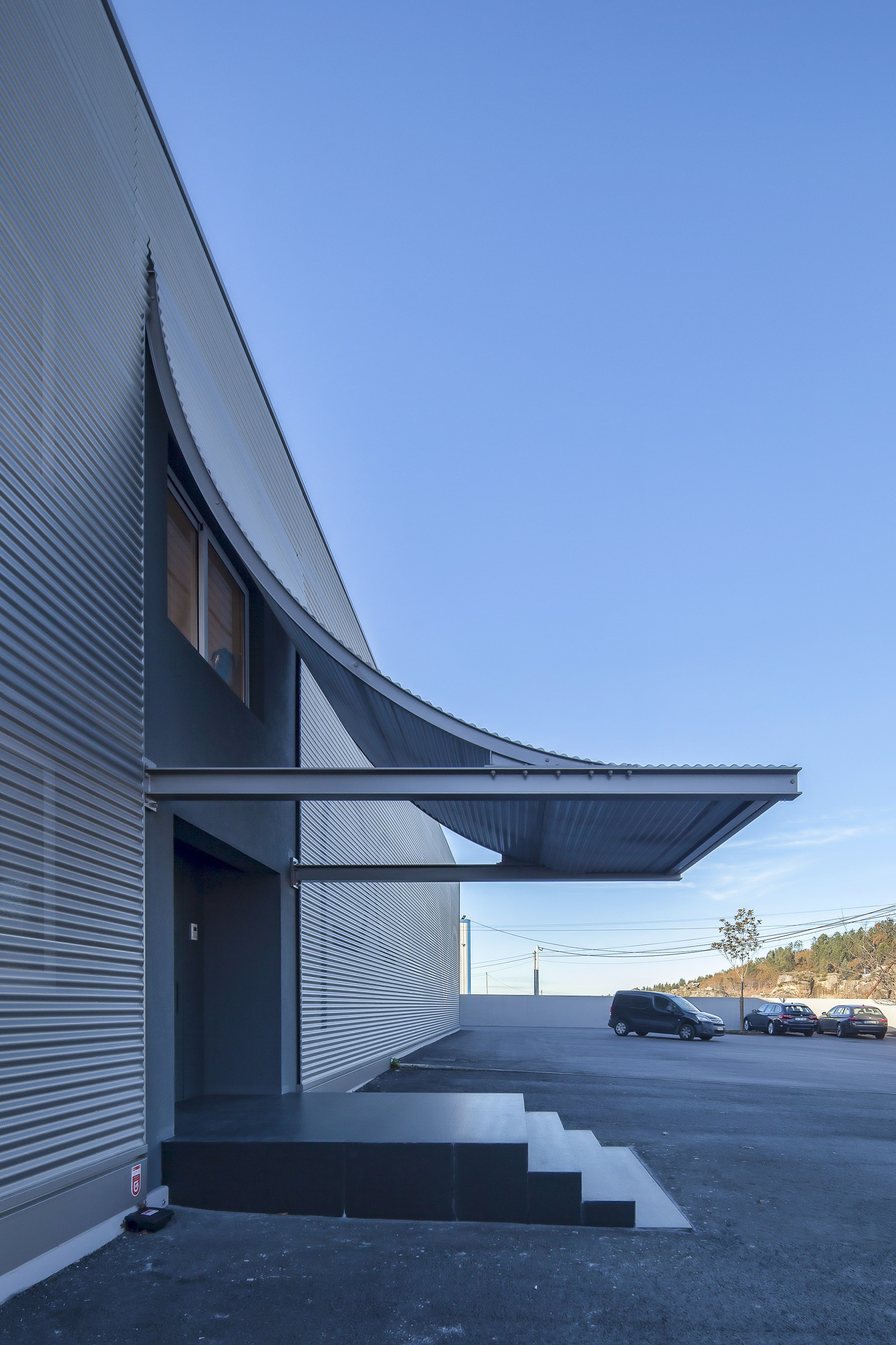 Side view of the sweeping canopy curving out from the cladding above black stone steps, with parked cars in the distance