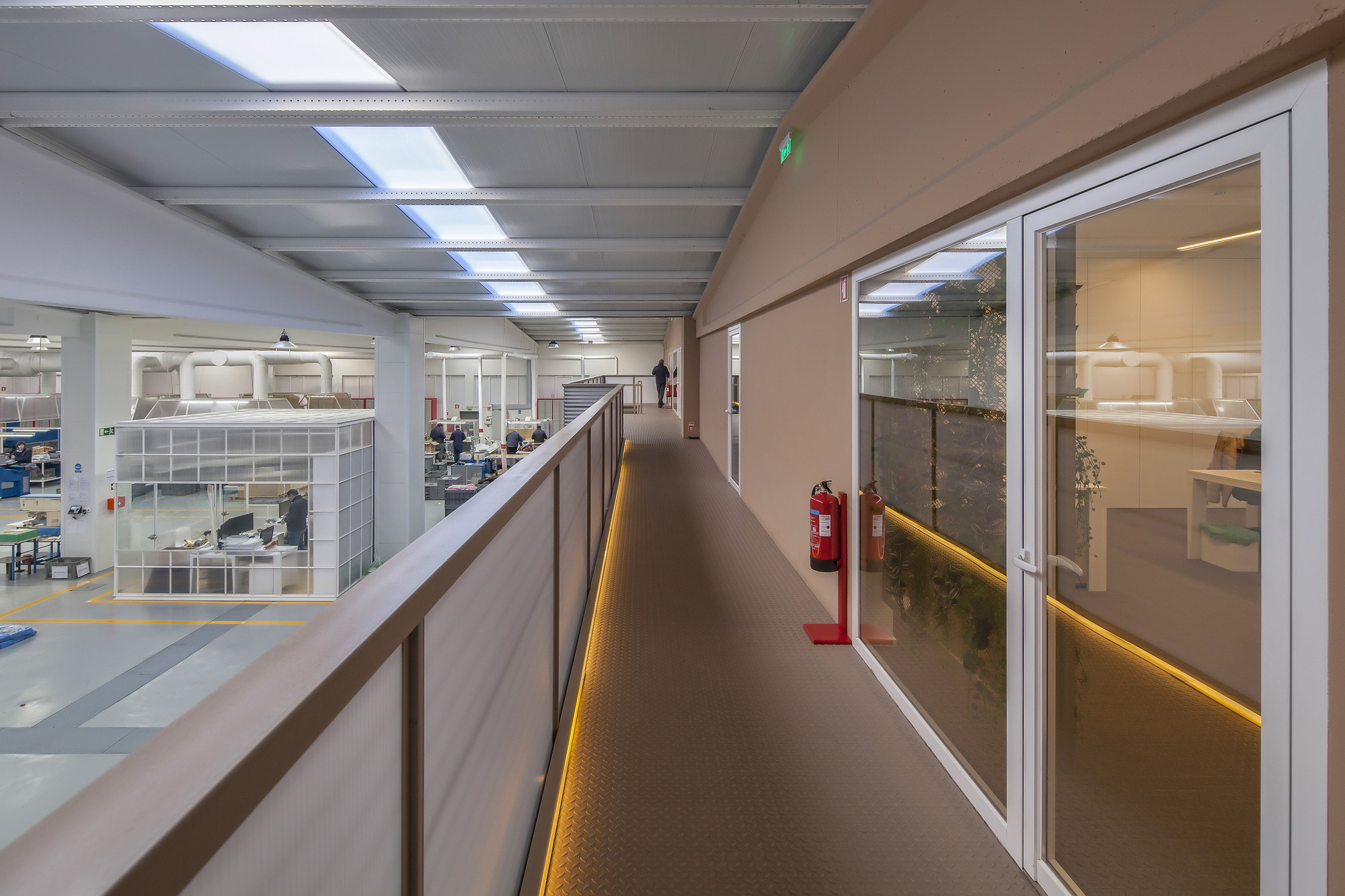 Raised walkway with a wooden-toned balustrade and glowing edge lights, overlooking the factory floor and skylights above