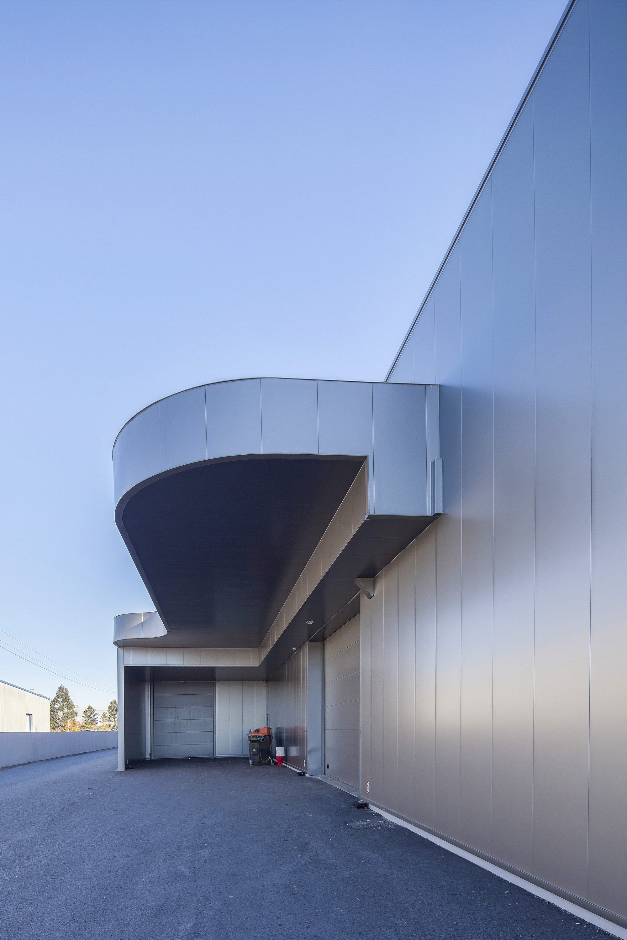 Sweeping underside of the curved canopy projecting from a long grey panel wall, seen against a clear blue sky