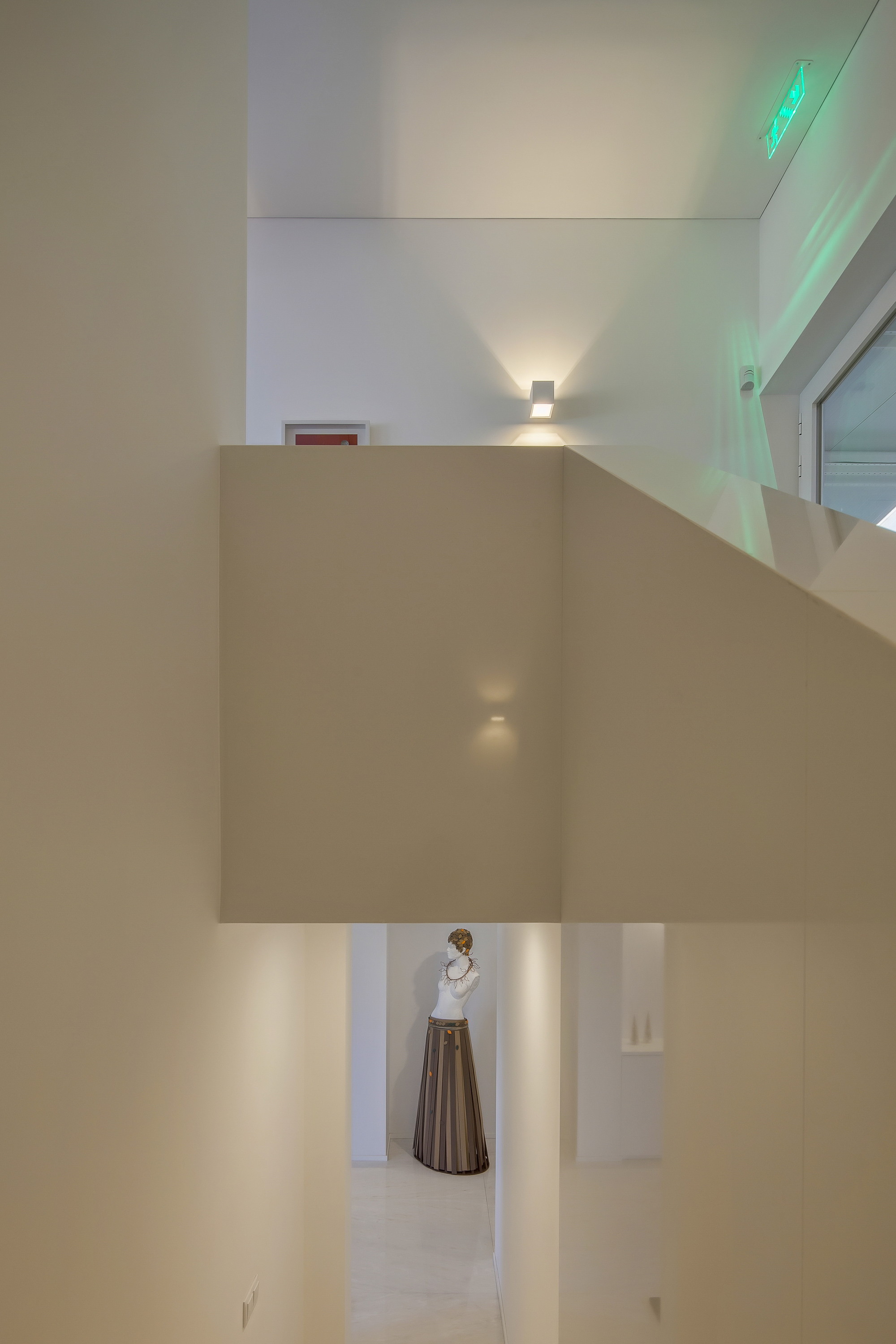 Narrow white stairwell opening onto a corridor where a dressed mannequin stands, lit by a wall sconce and green exit sign