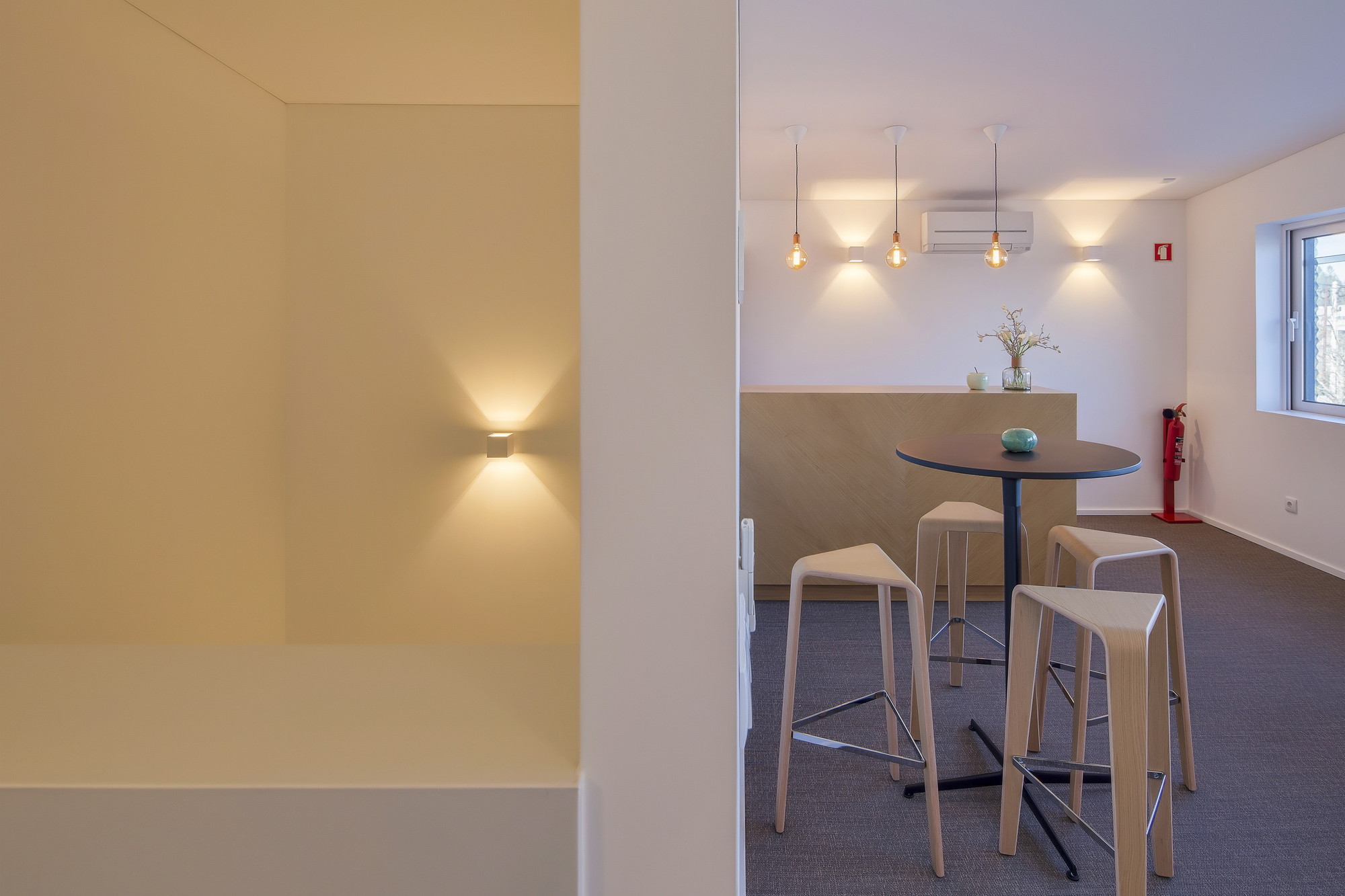 Break room with wooden bar stools, a round high table and pendant bulbs, seen past a wall with a lit sconce