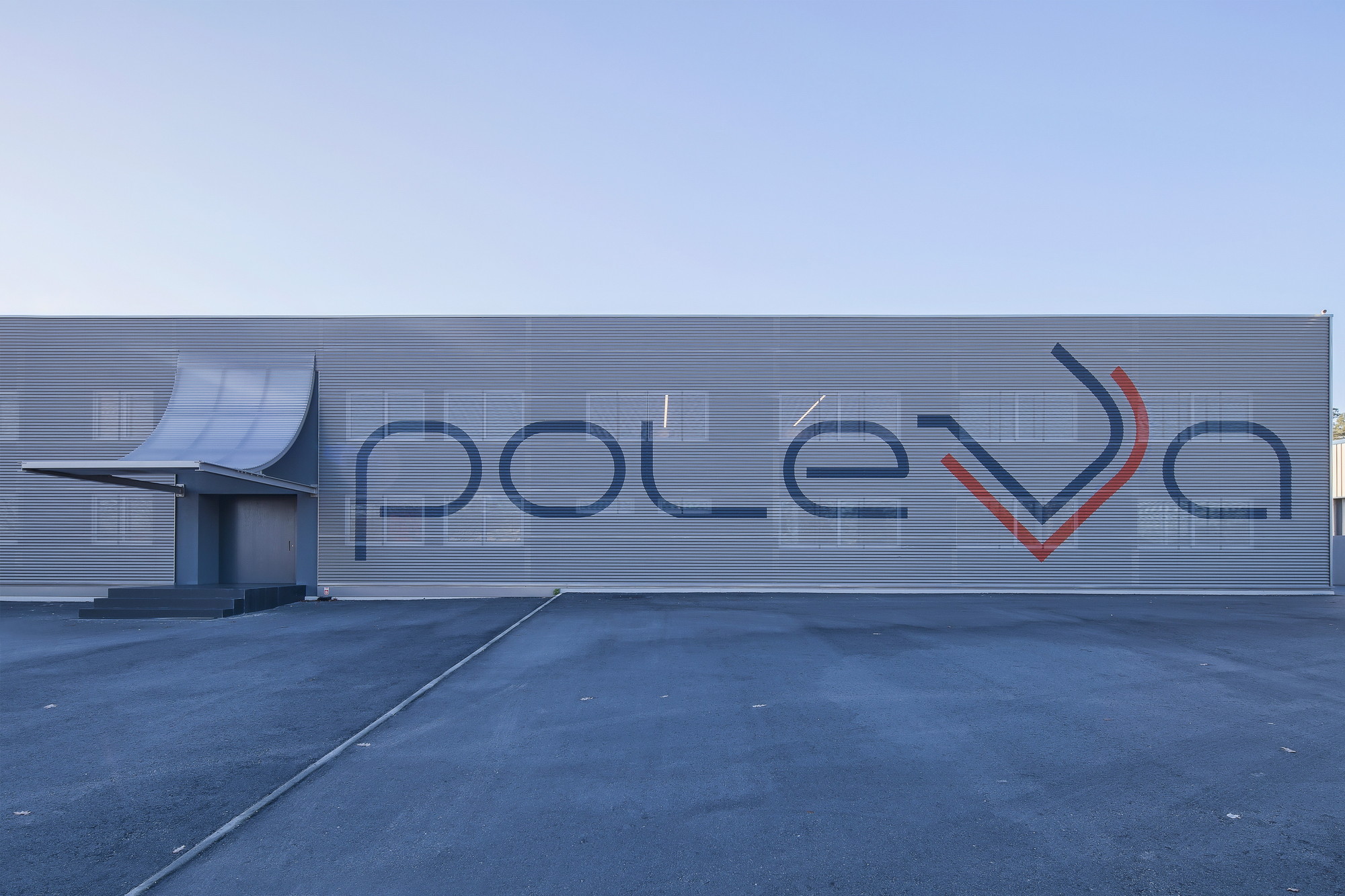 Frontal view of the long corrugated facade with large painted lettering and a small curved entrance canopy at left