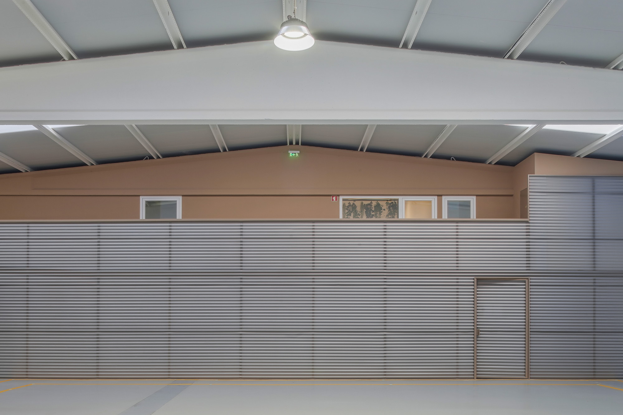 Interior of a pitched-roof hall with a corrugated grey metal wall, small high windows and a pendant lamp under cool overhead light