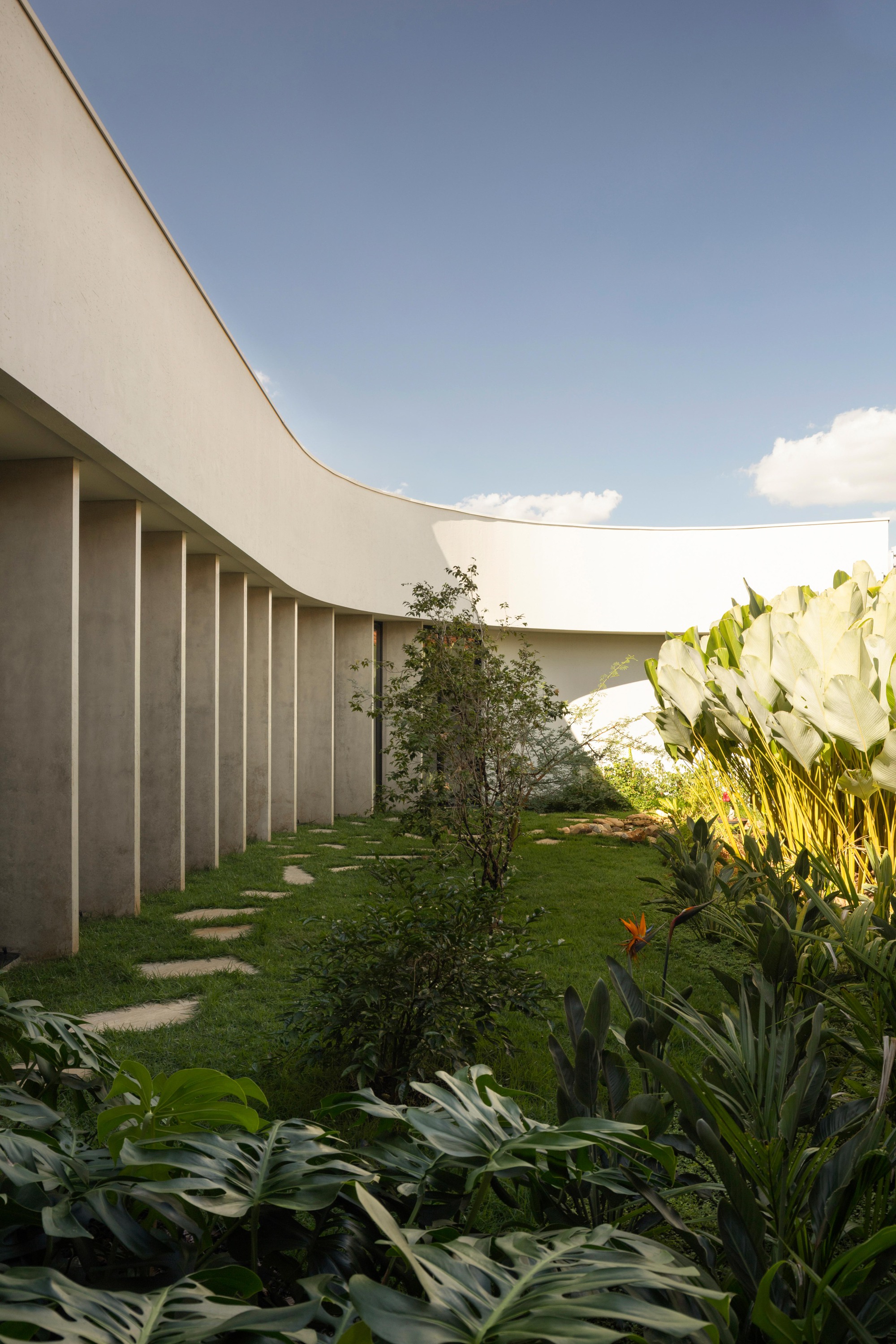 Curved colonnade of concrete fins beside a lawn with stepping stones, monstera leaves in the foreground under blue sky