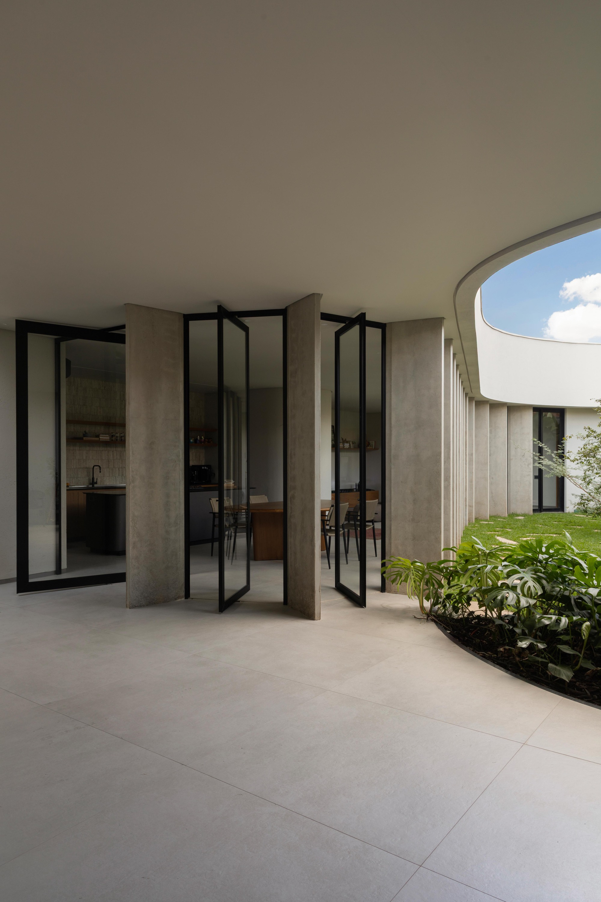 Pivoting black-framed glass doors opening from a kitchen and dining room onto a paved terrace beside a curved planting bed