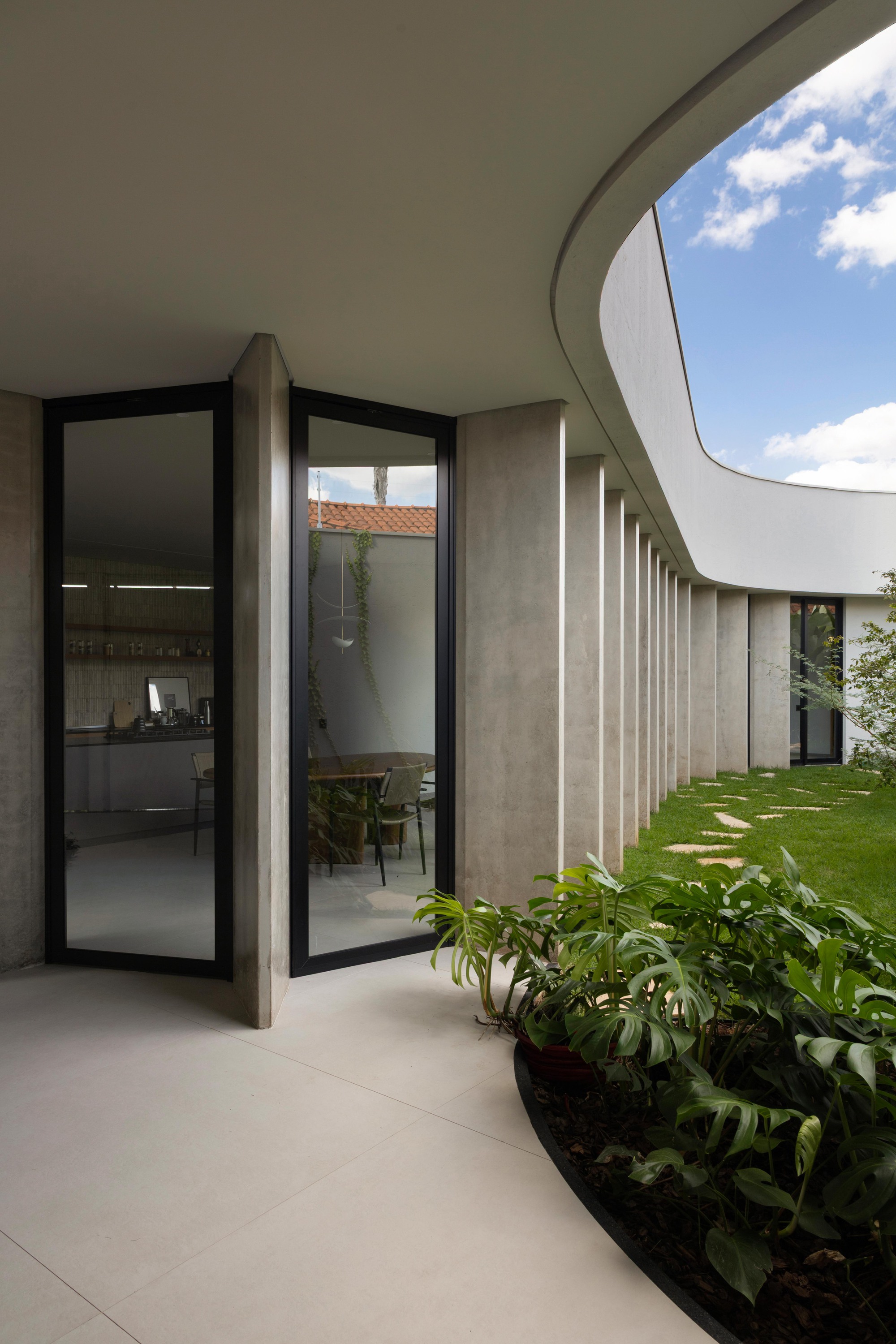 Corner of glazed doors between concrete piers, with a curved planter of monstera leaves and a courtyard lawn beyond