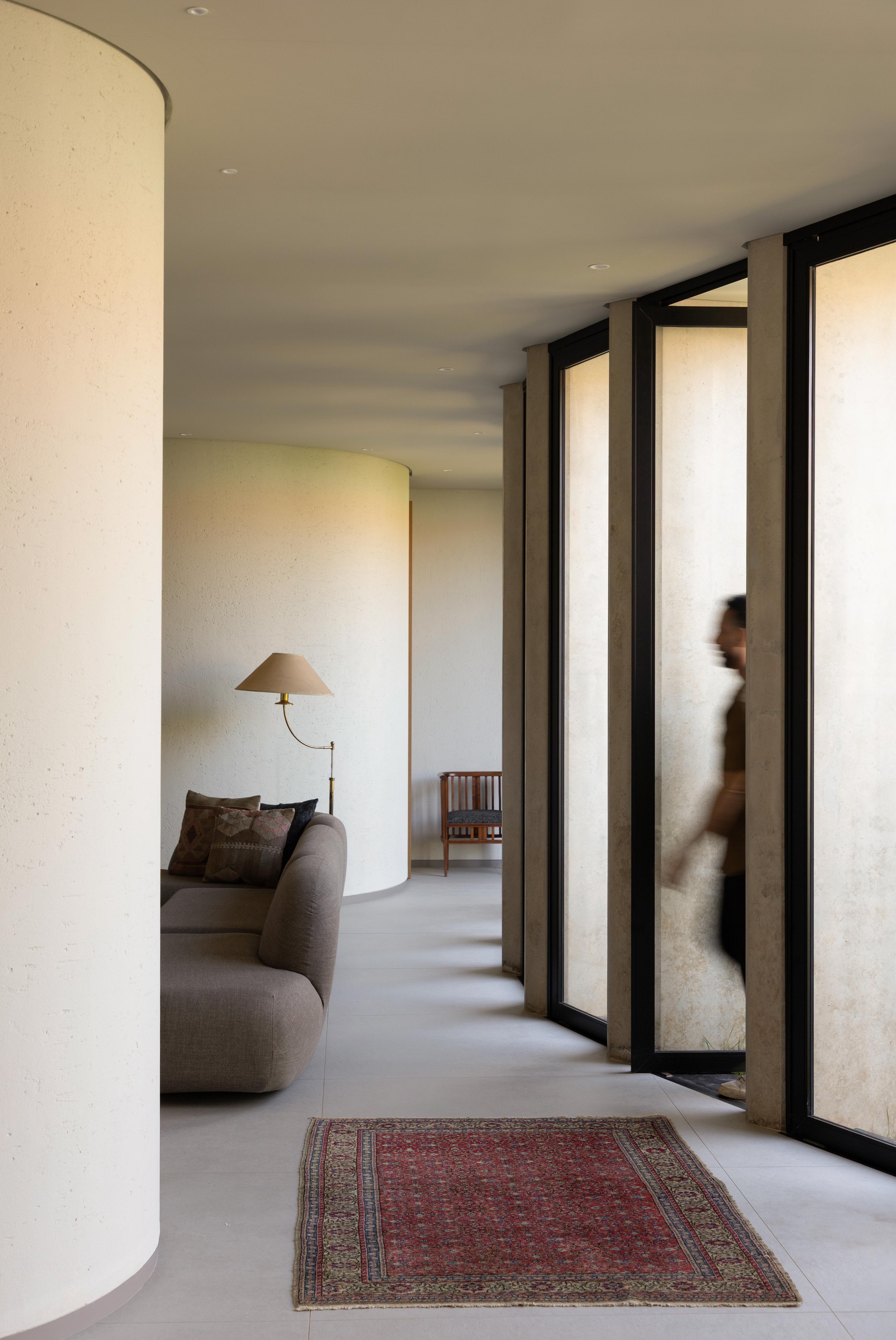 Interior corridor with a curved plaster column, a sofa, floor lamp and red rug, as a blurred figure walks past tall glazing