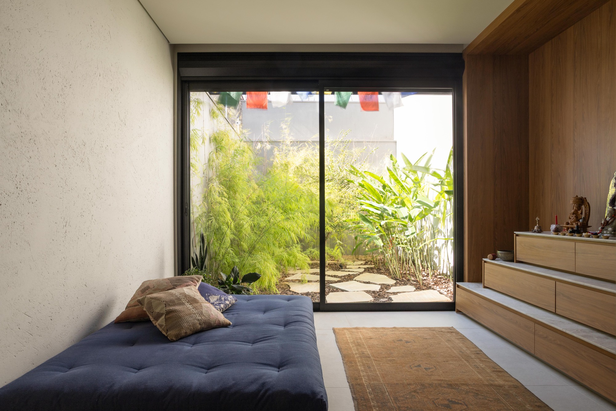 Low navy daybed facing a sliding glass door that opens onto a bamboo courtyard, with prayer flags and stepped wood shelves beside it