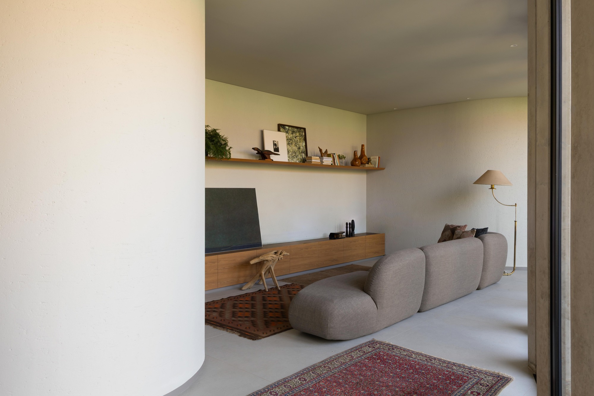 Curved plaster wall beside a living area with a rounded grey sofa, long oak console and shelf, and layered rugs in soft daylight