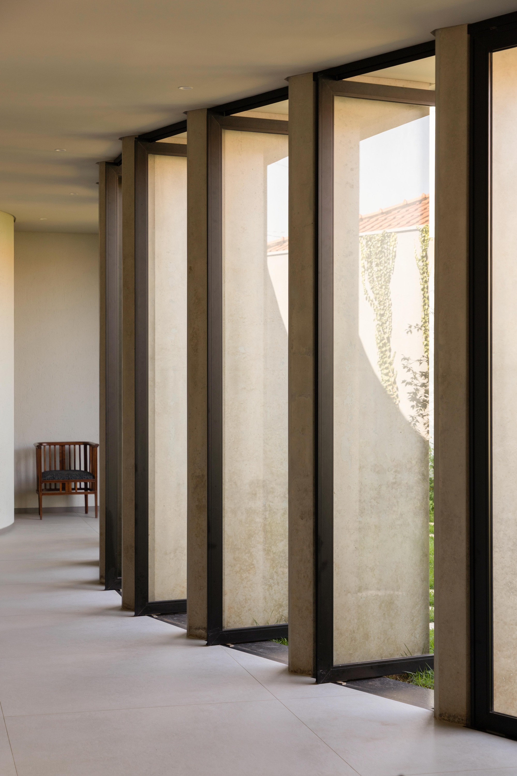 Row of tall pivoting glass panels framed in black steel and concrete fins, with a wooden chair at the end of the corridor