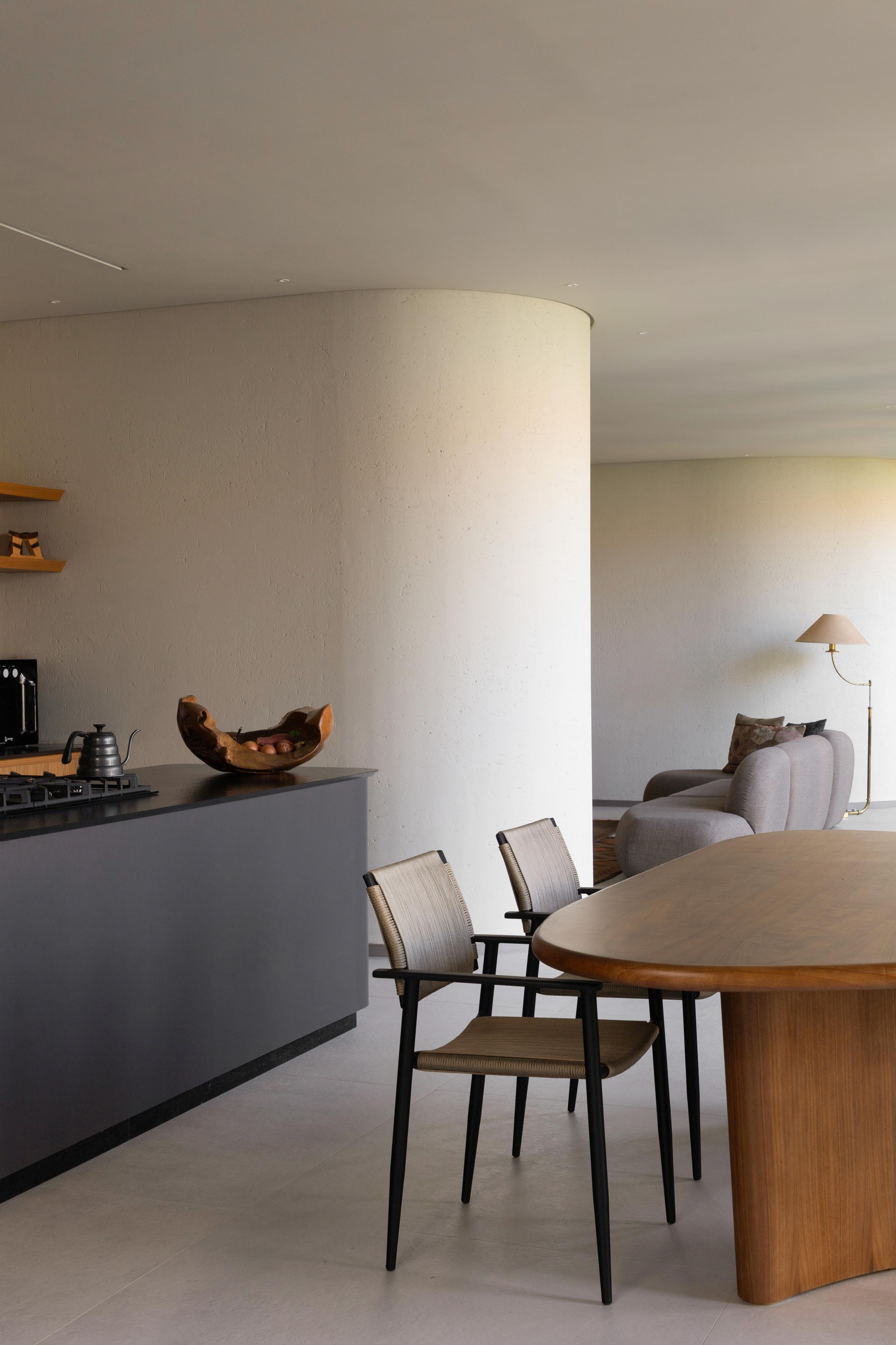 Dining area with an oval timber table and slim chairs, a dark kitchen island and a curved plaster wall behind