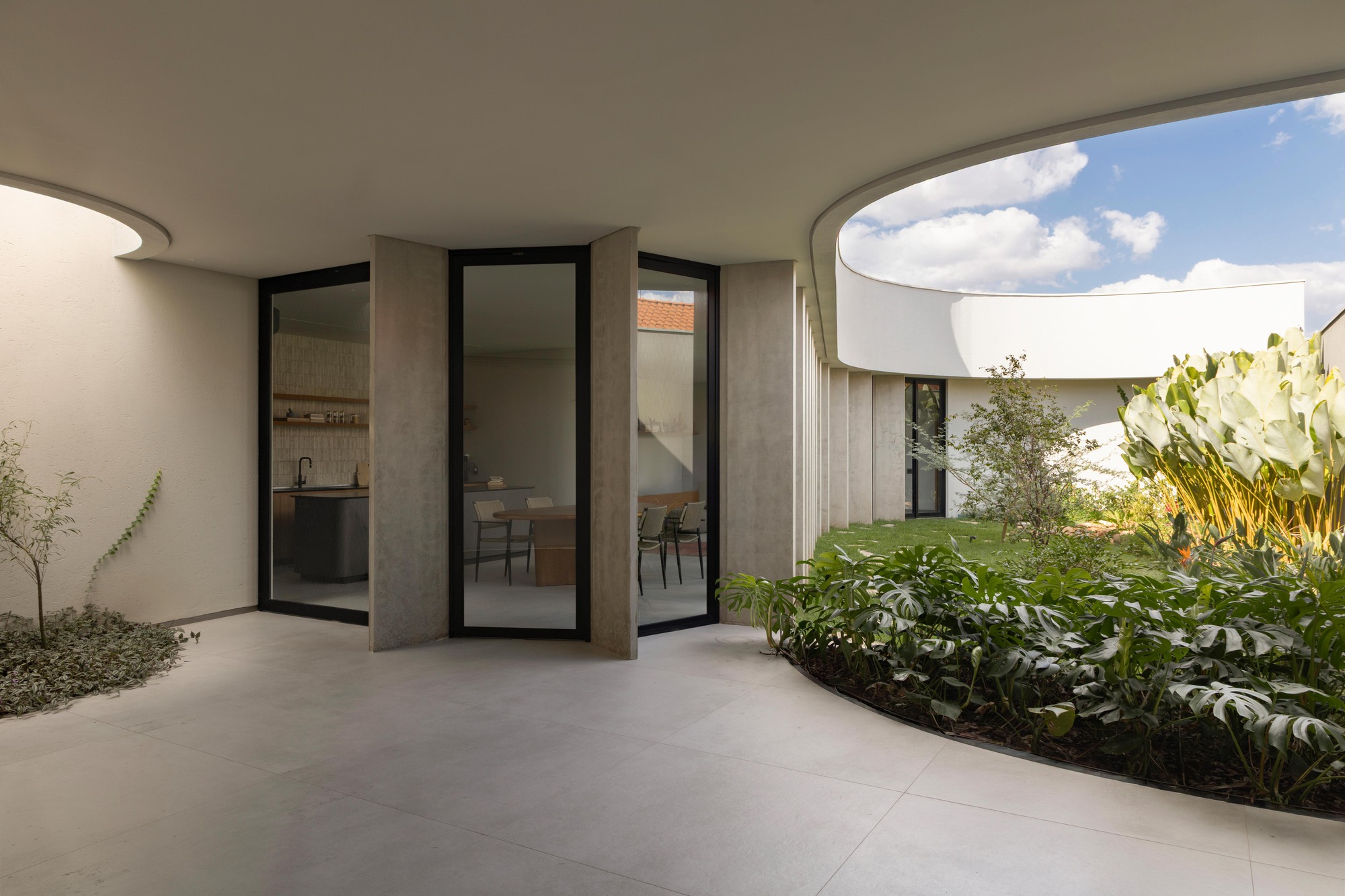Covered terrace with pivoting glass doors facing a curved planted bed of monstera and a round roof opening under a cloudy blue sky
