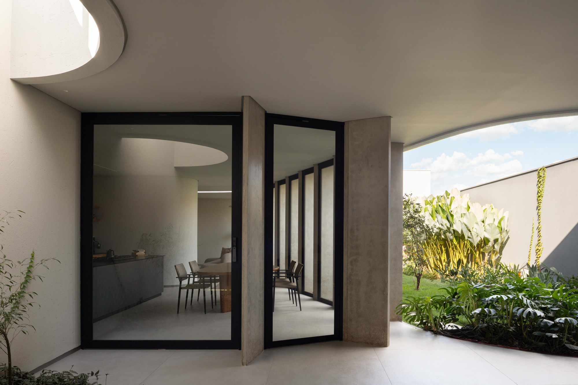 Pivoting glass door between concrete fins, reflecting the dining table, beside a planted courtyard under a curved overhang in afternoon sun