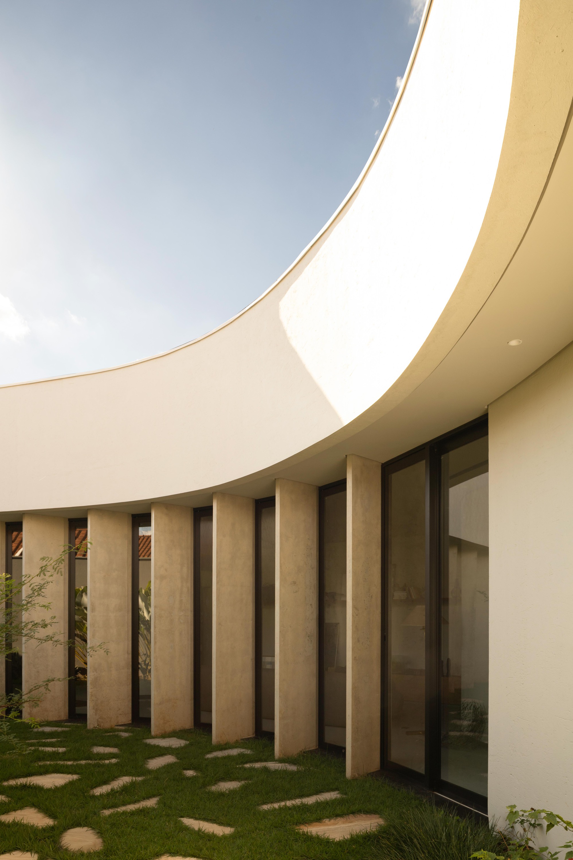 Curved white roof edge above tall stone fins and glazing, with stepping stones set in grass under a clear blue sky