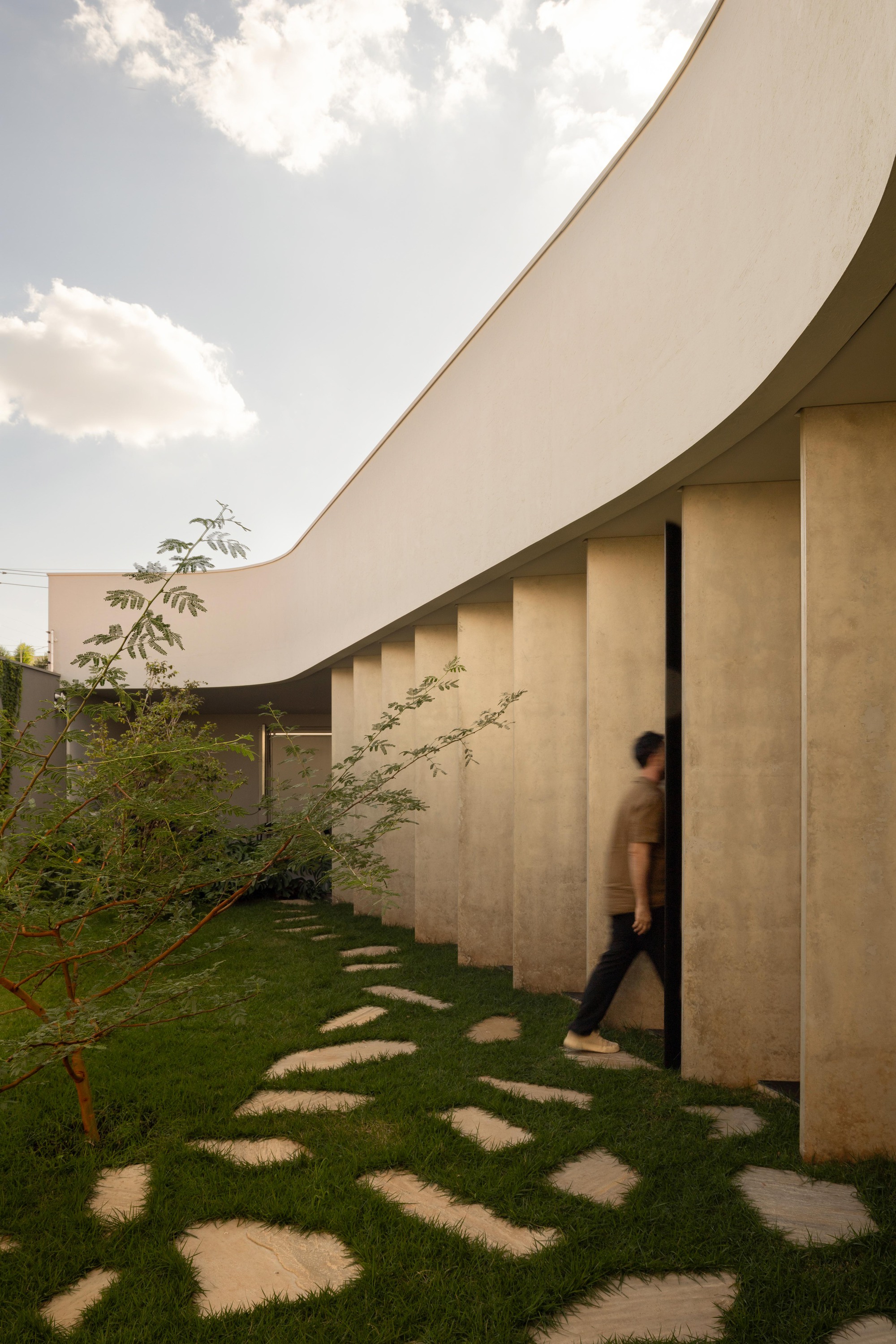 Person walking past rows of stone fins beneath the curving white roof, with stepping stones in grass and a young tree
