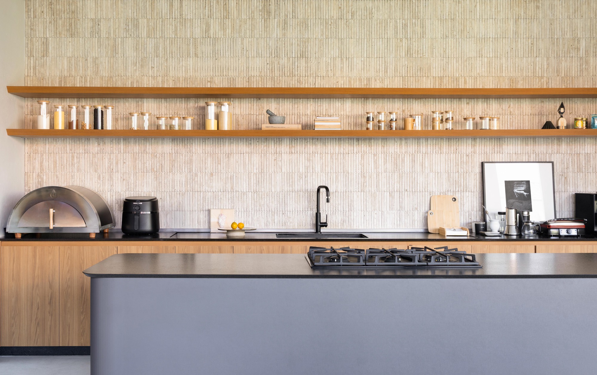 Kitchen with two open timber shelves of glass jars above a black sink, with a gas hob on the grey island