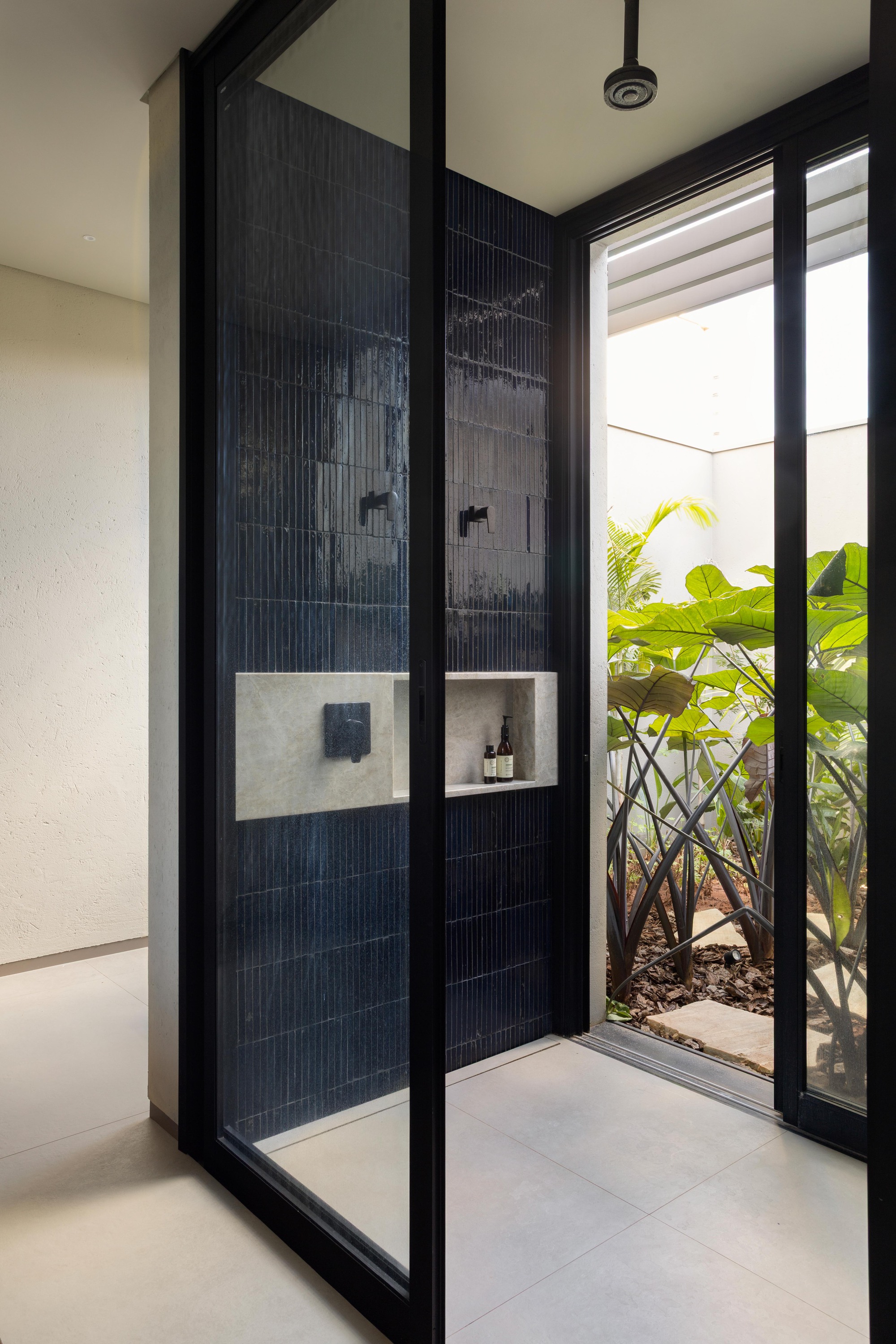 Glass-enclosed shower with dark blue vertical tiles and a stone niche, opening onto a planted light well with broad-leaved plants