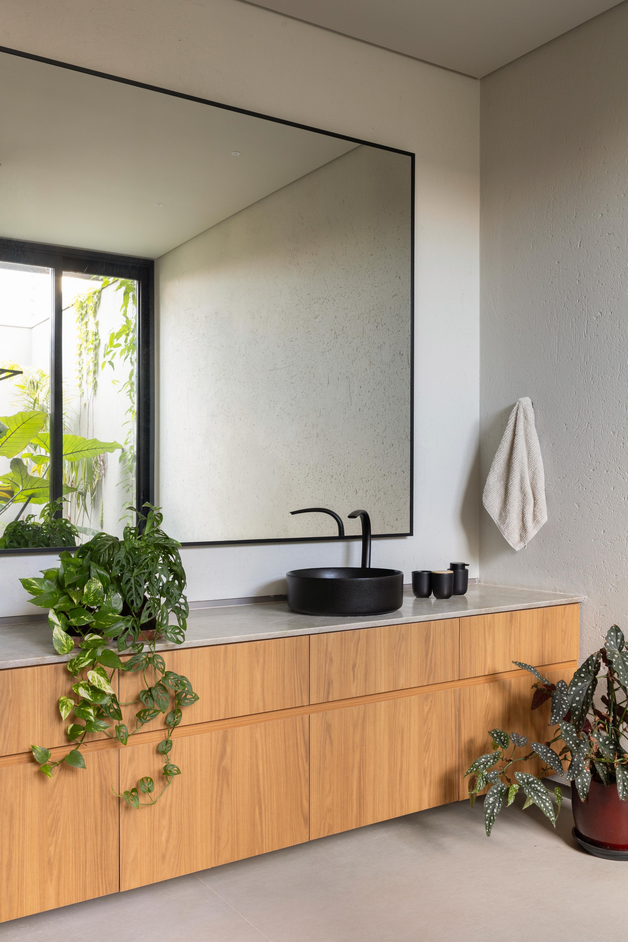 Oak vanity with a black round basin beneath a large framed mirror, trailing pothos and a begonia in soft daylight