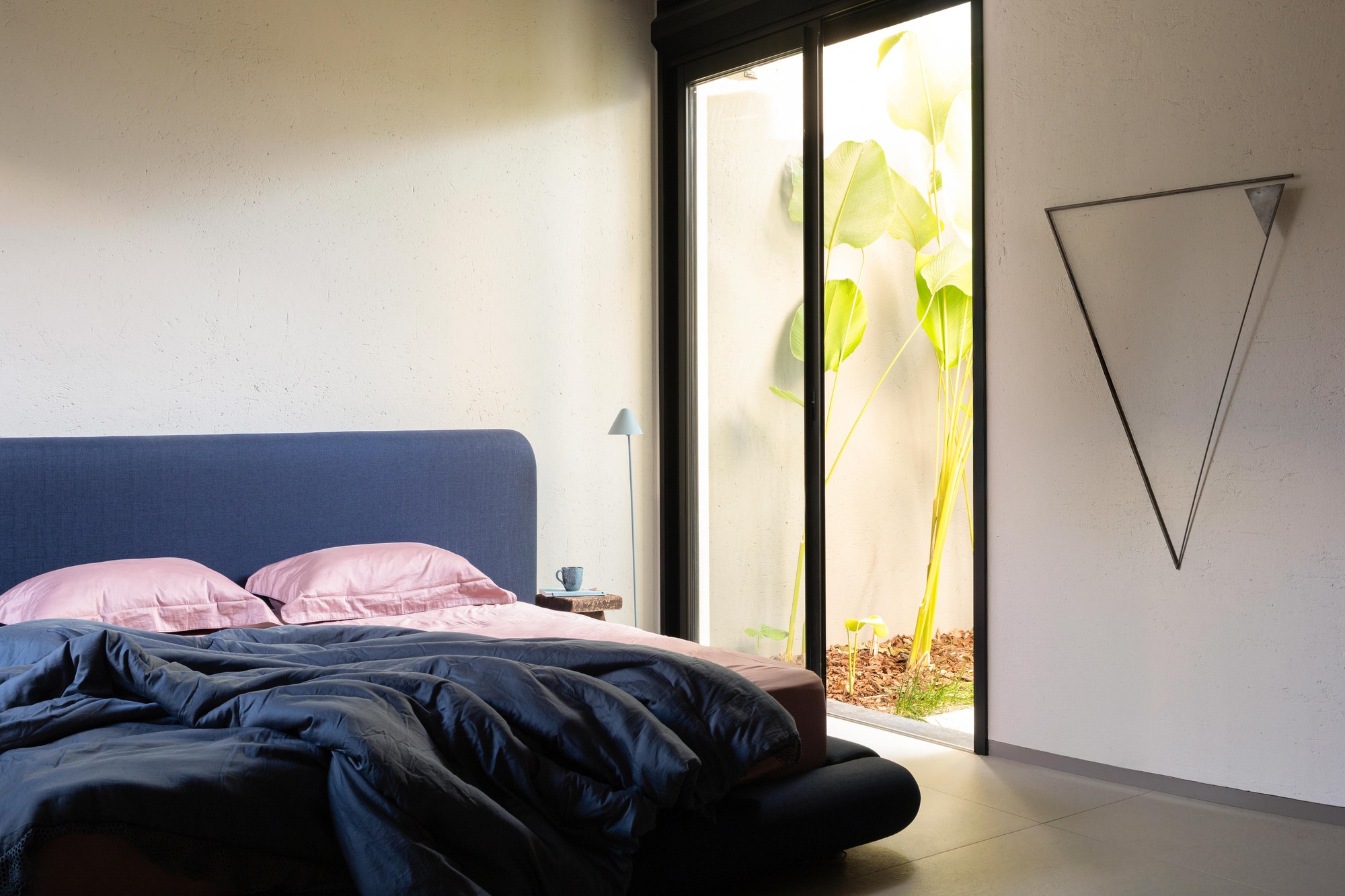 Bedroom with a blue upholstered headboard and rumpled bedding beside a glass door onto a sunlit courtyard of banana leaves