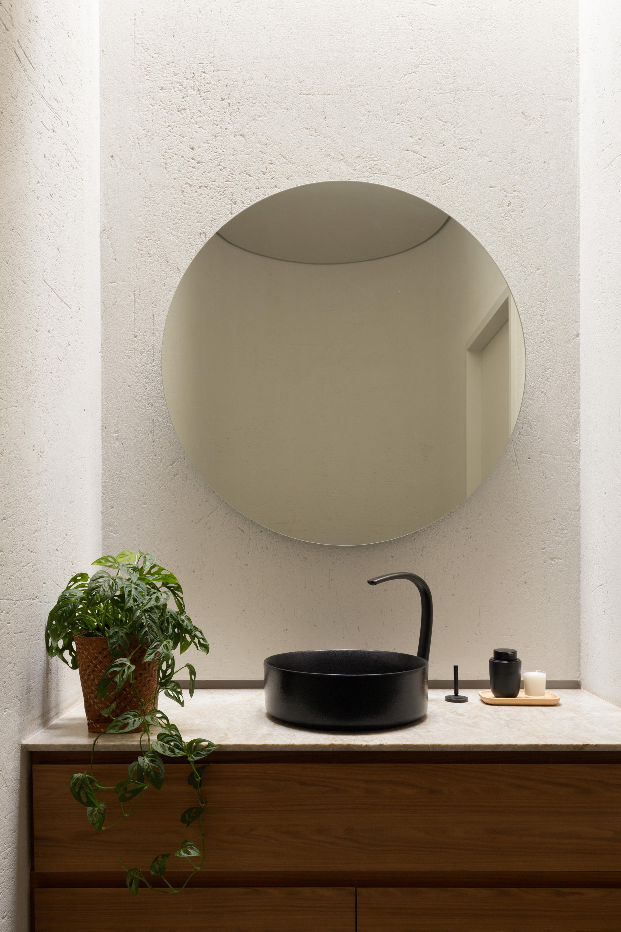 Round mirror above a black vessel basin on a timber vanity, with a trailing plant against textured plaster walls