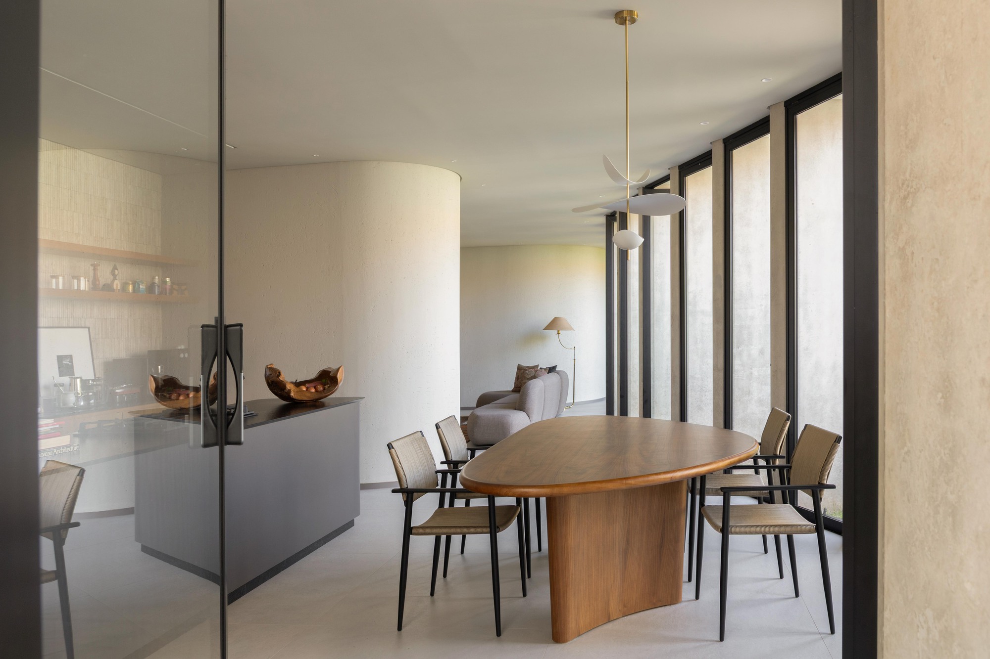 Dining room with an oval timber table and woven chairs, curved plaster wall and tall slotted windows letting in afternoon light