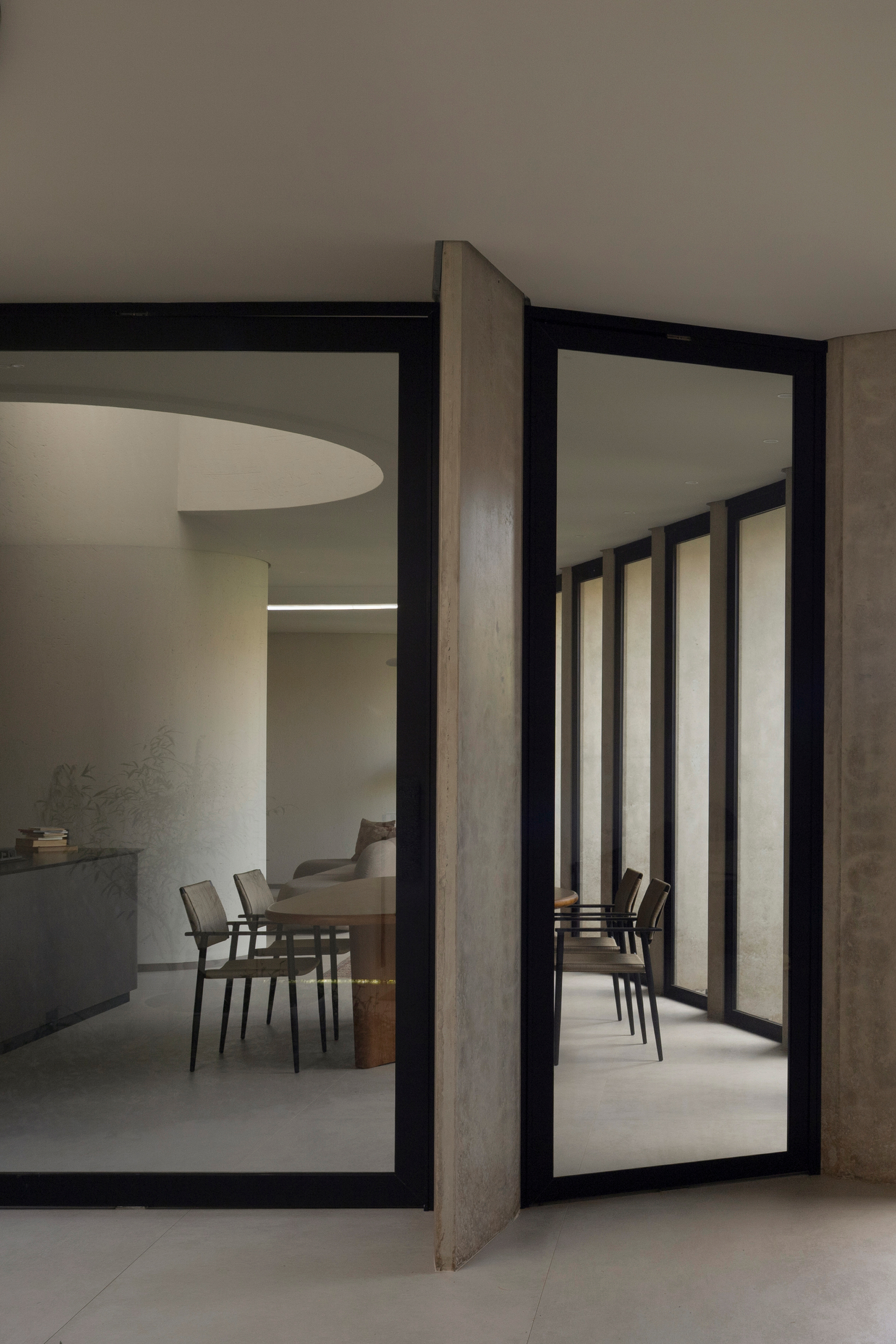 Pivoting stone door panel between black-framed glass, revealing a curved dining space and chairs beyond in muted daylight