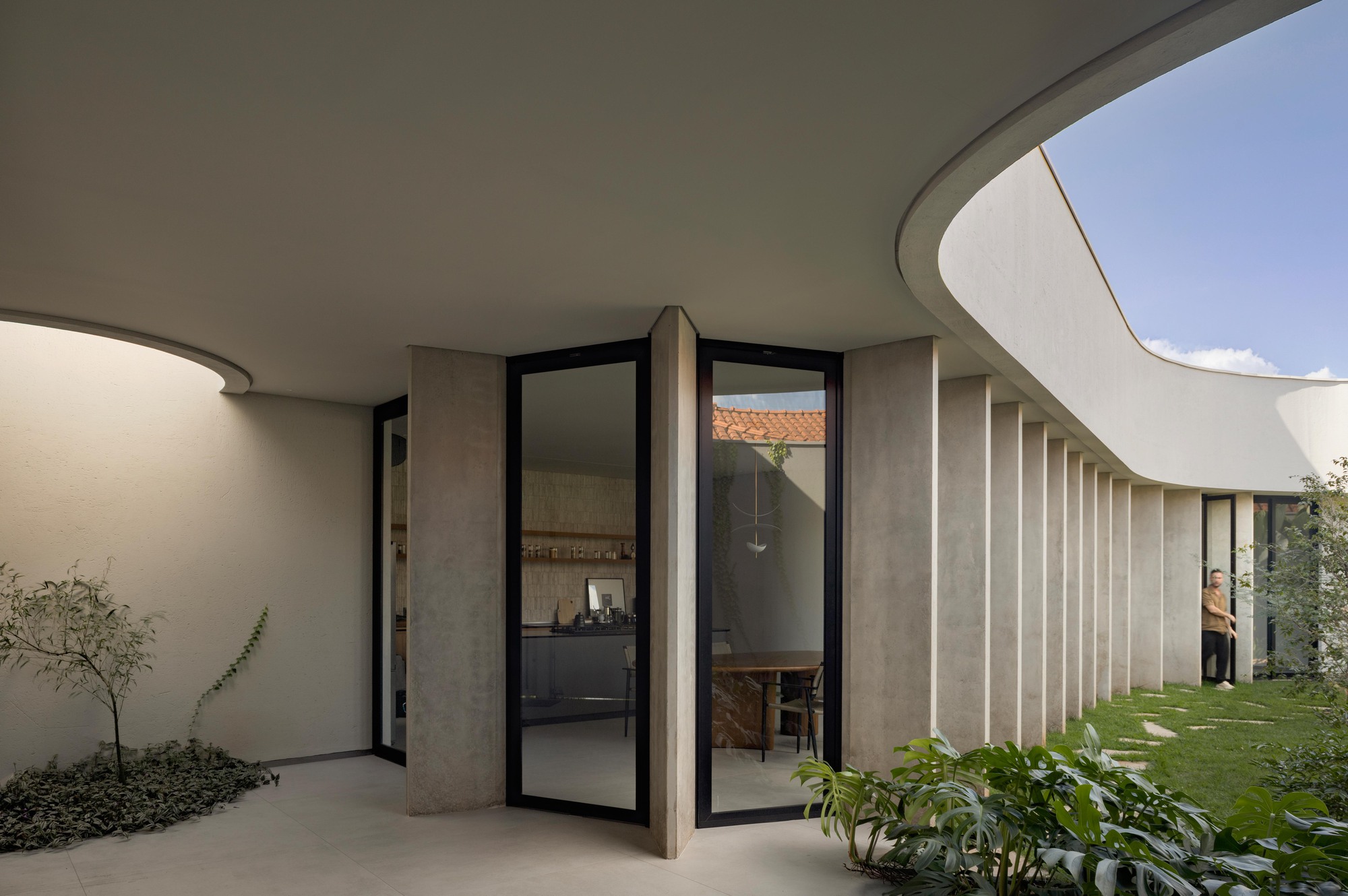 Covered walkway with concrete fins and black-framed glass doors curving around a lawn garden, a man standing in the far doorway under blue sky