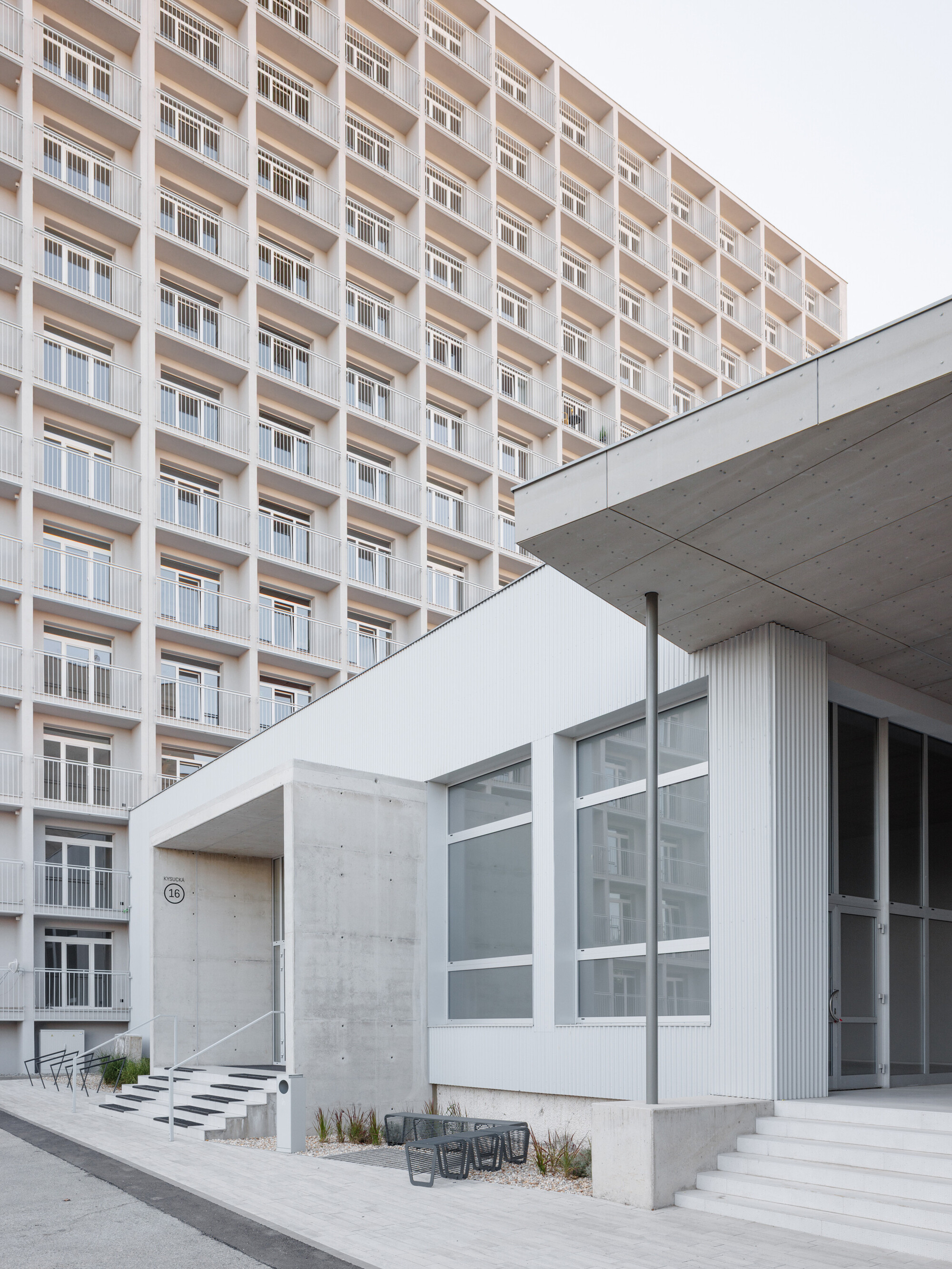 Concrete entrance canopy and corrugated white volume in front of the balconied tower, with steps and a bench in daylight