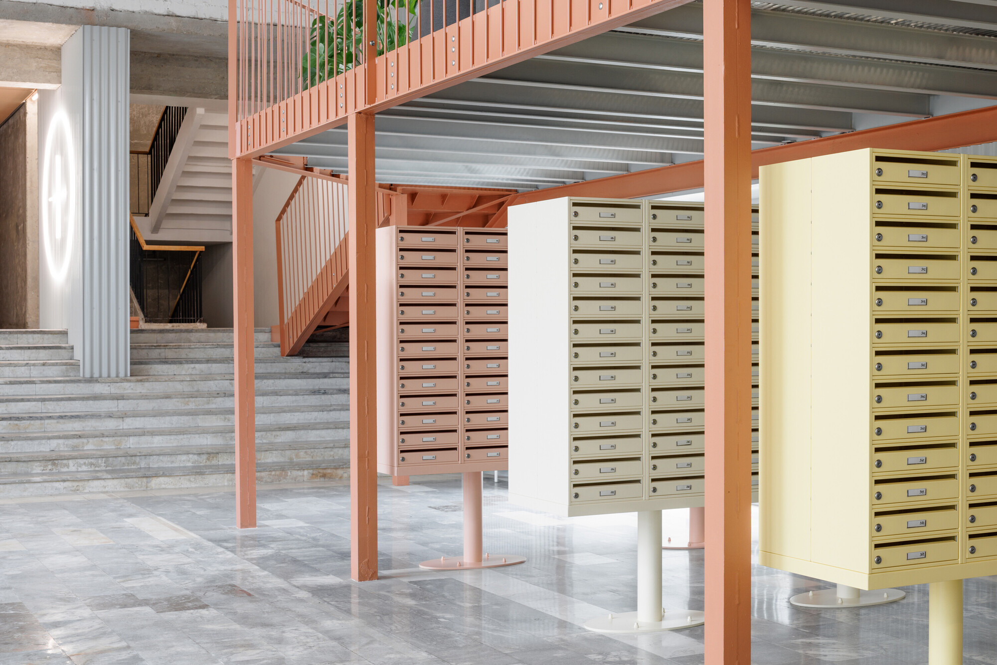 Pastel mailbox cabinets standing beneath a coral steel mezzanine, with wide stone steps rising behind on a polished floor