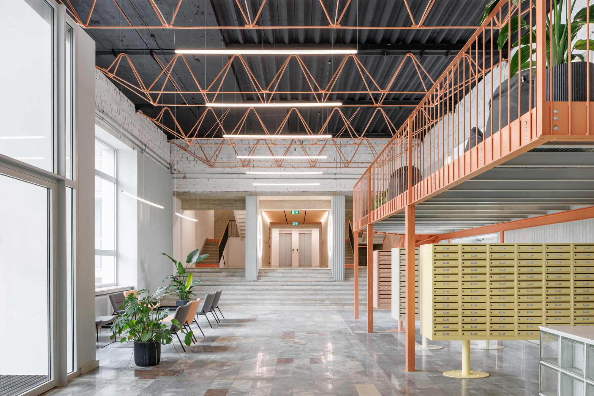 Lobby with coral steel truss ceiling, mezzanine and yellow mailboxes, with potted plants and chairs by a tall window