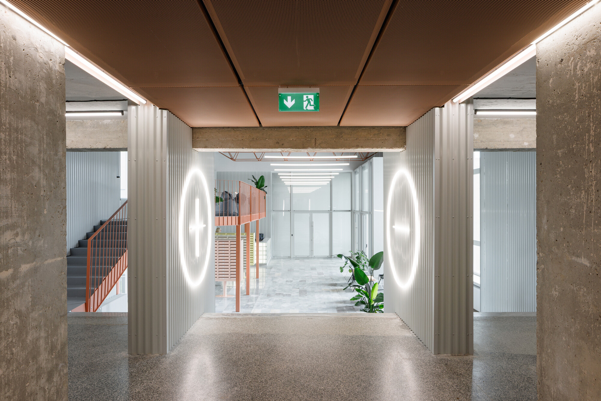 Concrete-framed passage with copper ceiling panels and glowing circular lights on corrugated walls, leading toward a plant-lit lobby