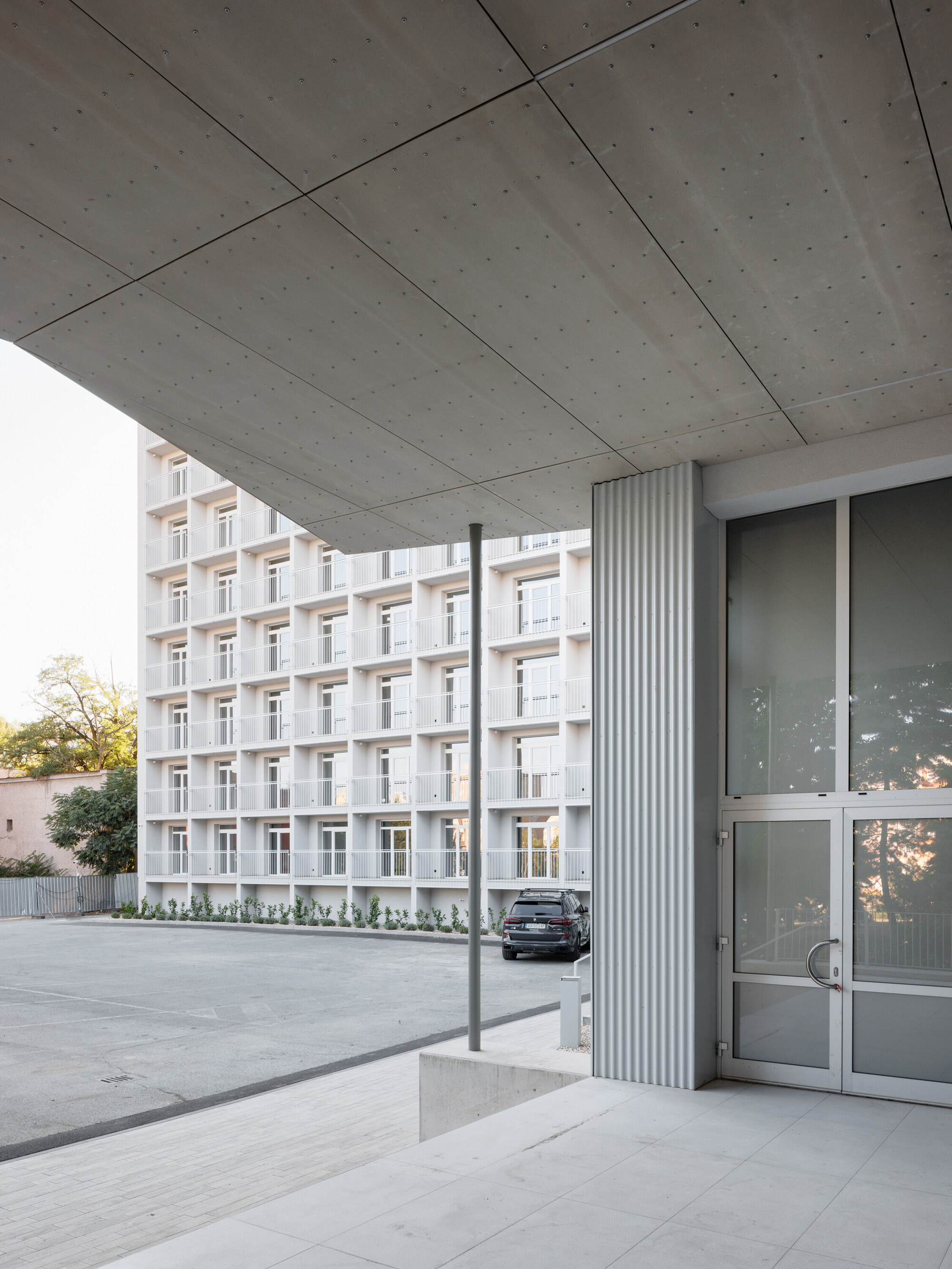 Concrete canopy soffit and corrugated metal column framing a gridded white residential block, with a parked car in the sunlit courtyard