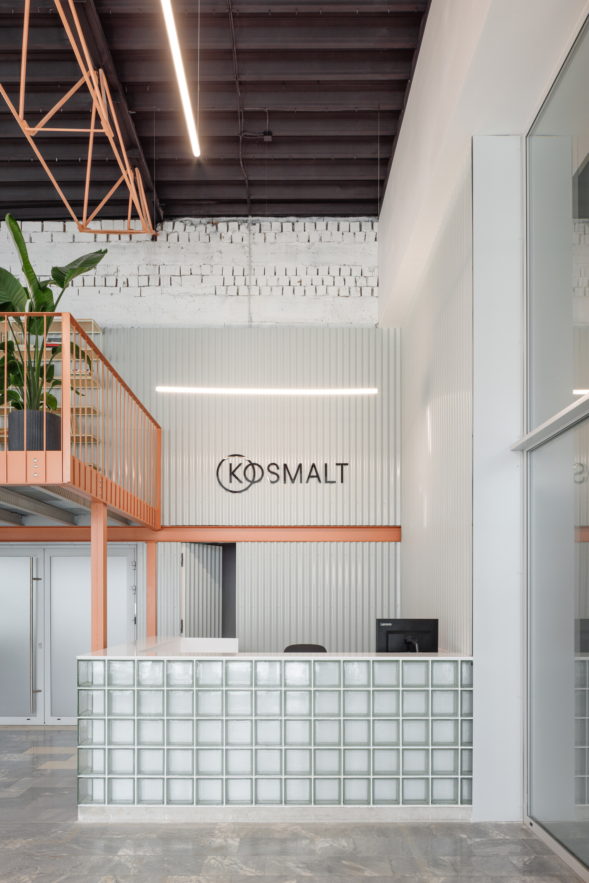 Reception desk clad in glass blocks below a coral mezzanine and corrugated metal wall with a wall-mounted sign