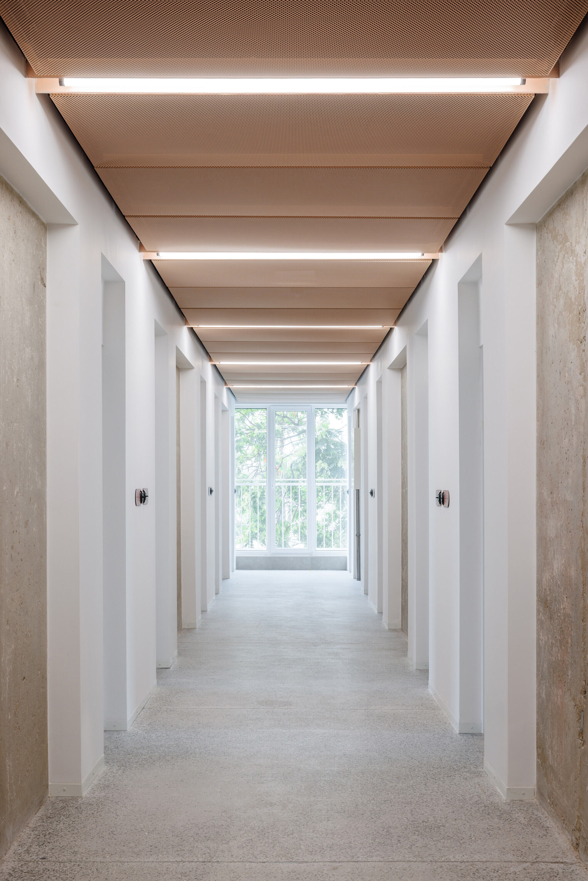 Corridor of white door recesses and bare concrete walls leading to a bright window, under a perforated copper ceiling with light strips