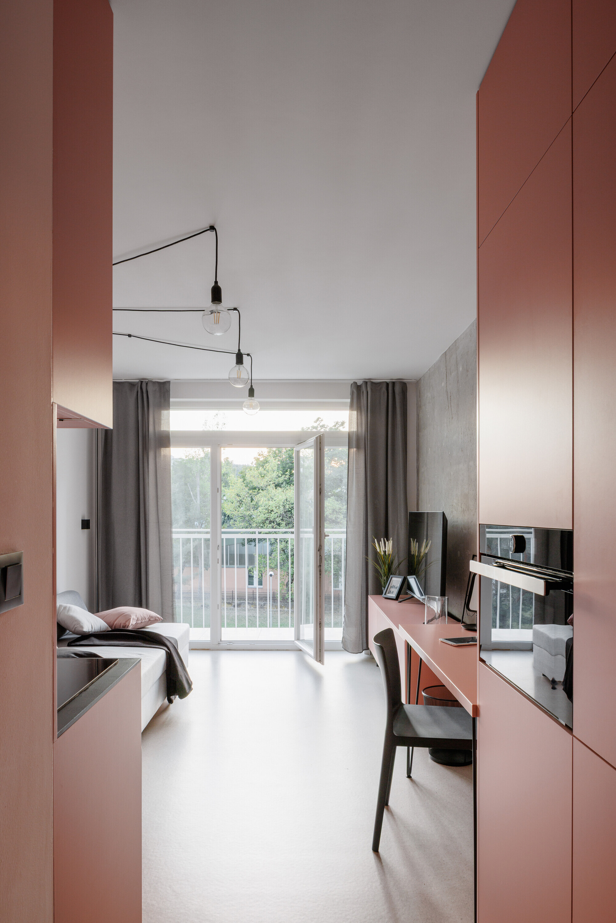Narrow studio apartment between salmon-pink cabinetry, with a desk, bed and open balcony door framing green treetops in daylight