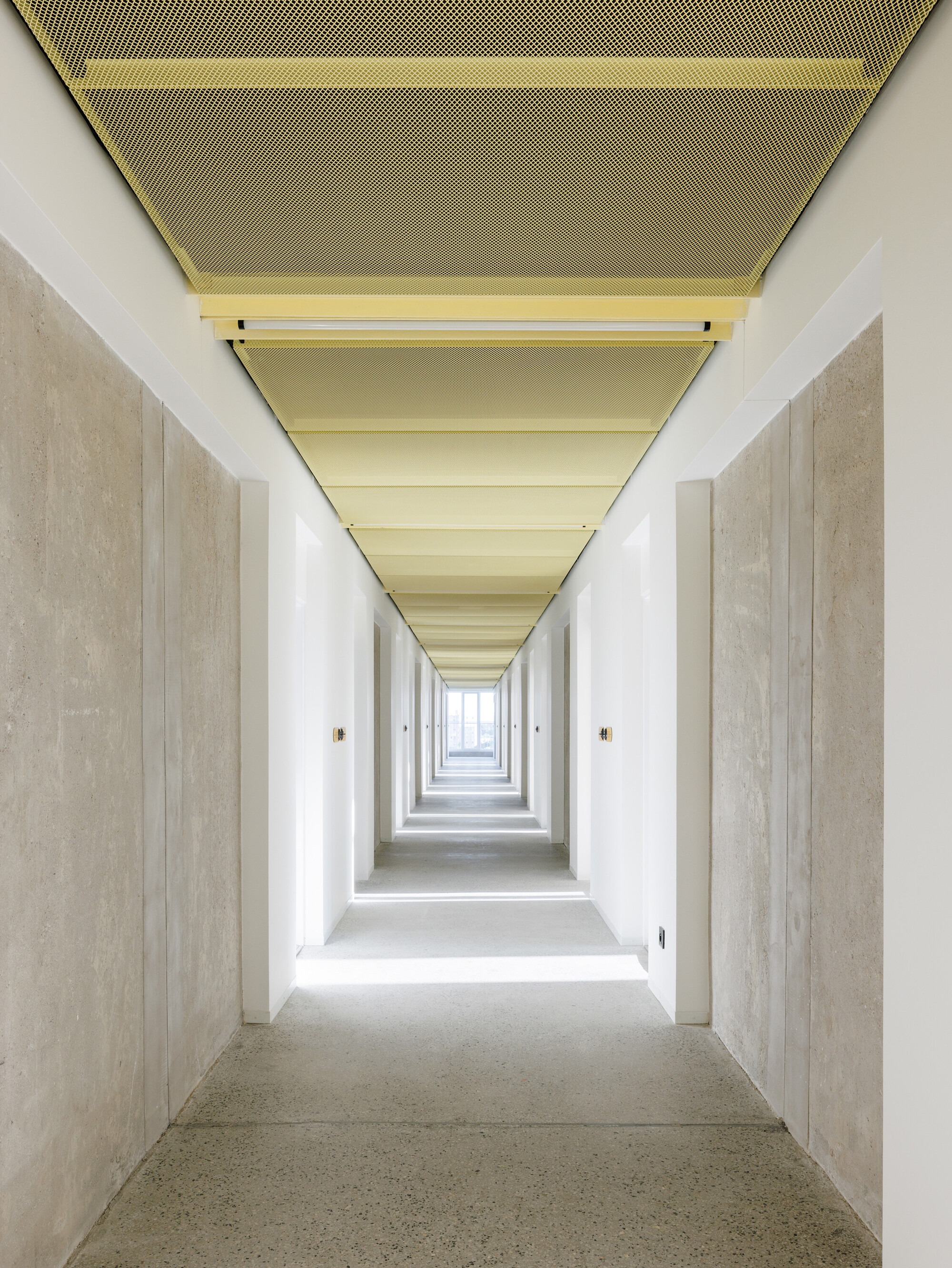 Long corridor under a yellow perforated metal ceiling, with concrete walls and daylight spilling across the terrazzo floor