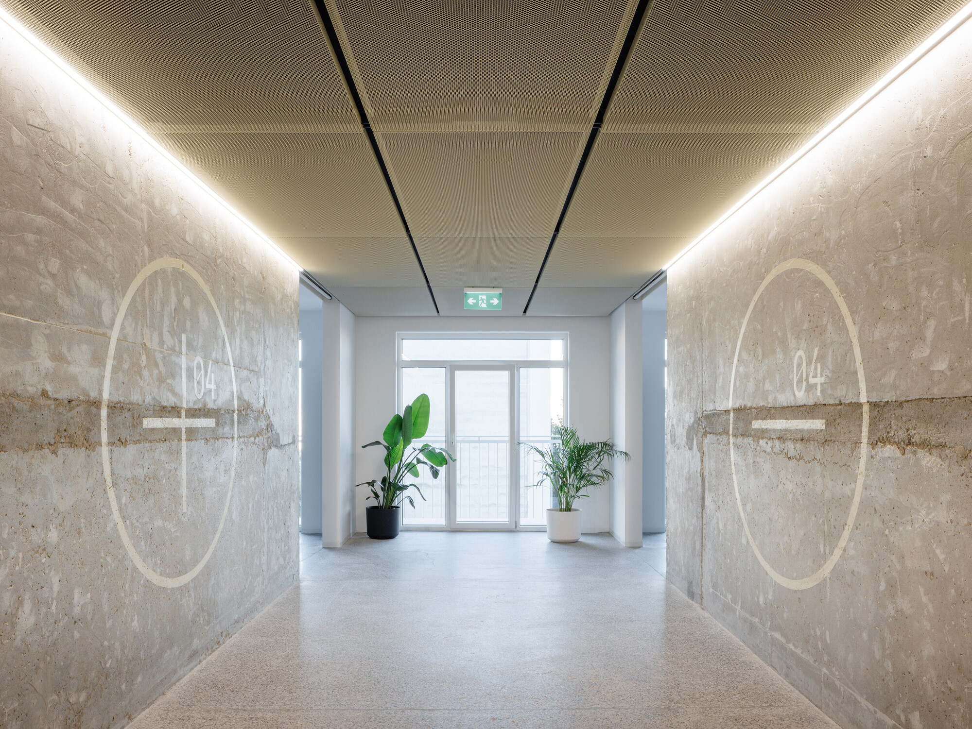 Corridor with two concrete walls marked by painted circles and number 04, lit by linear strips above and potted plants at the end