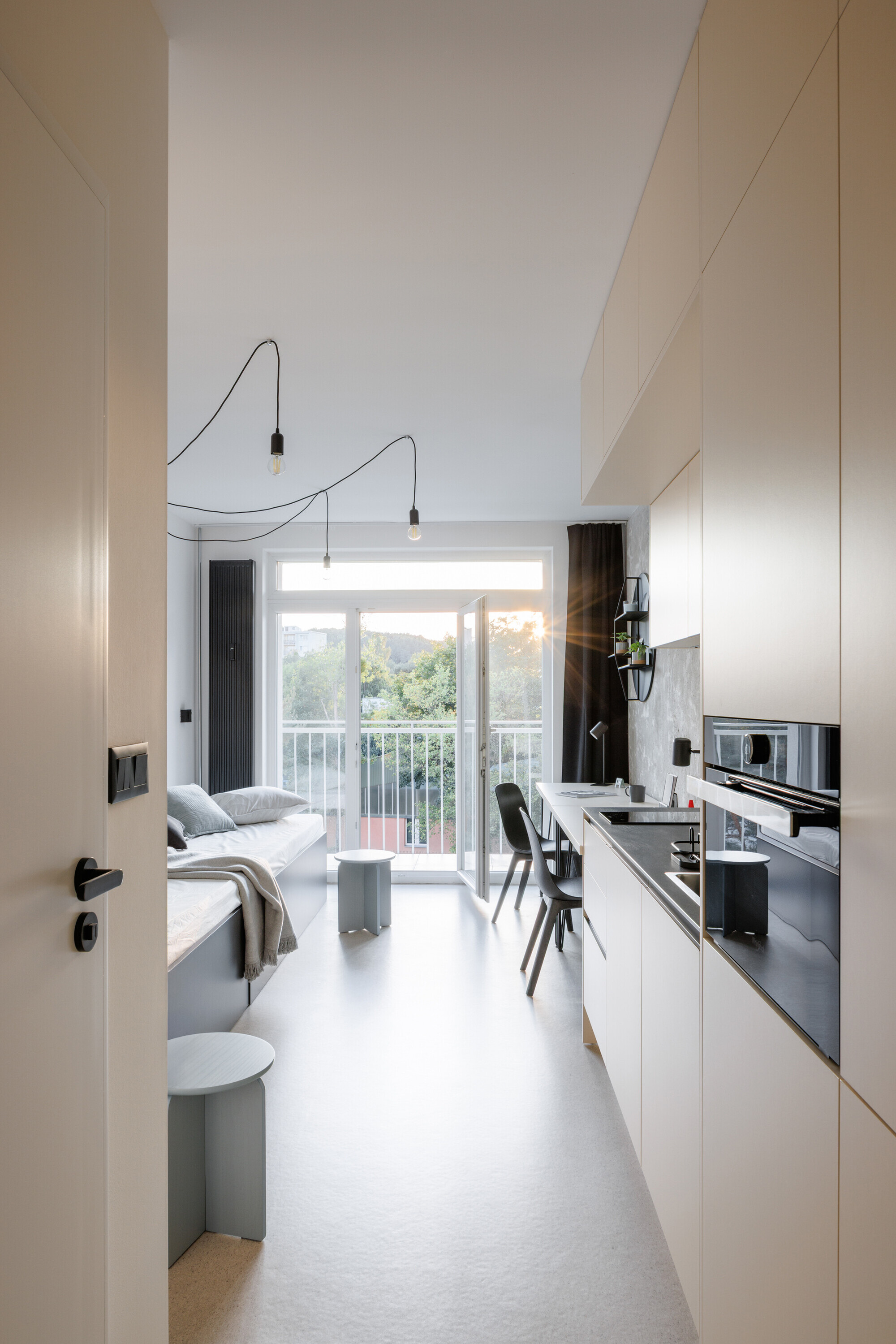 Narrow studio apartment seen from the entry door, with a kitchen run, daybed and open balcony door catching low sun