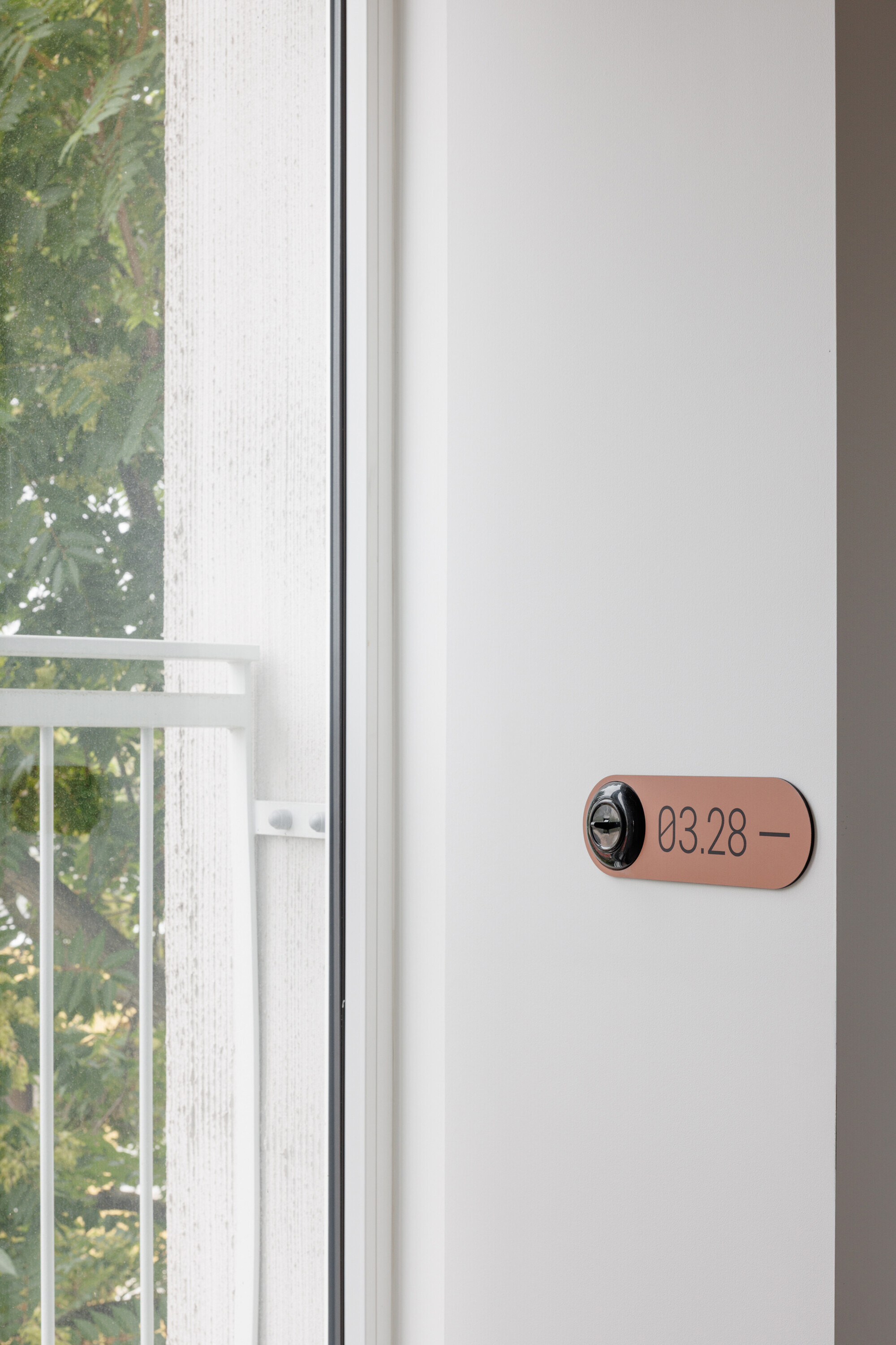 Copper-toned room number plate with a lock cylinder on a white wall beside a balcony door and leafy tree