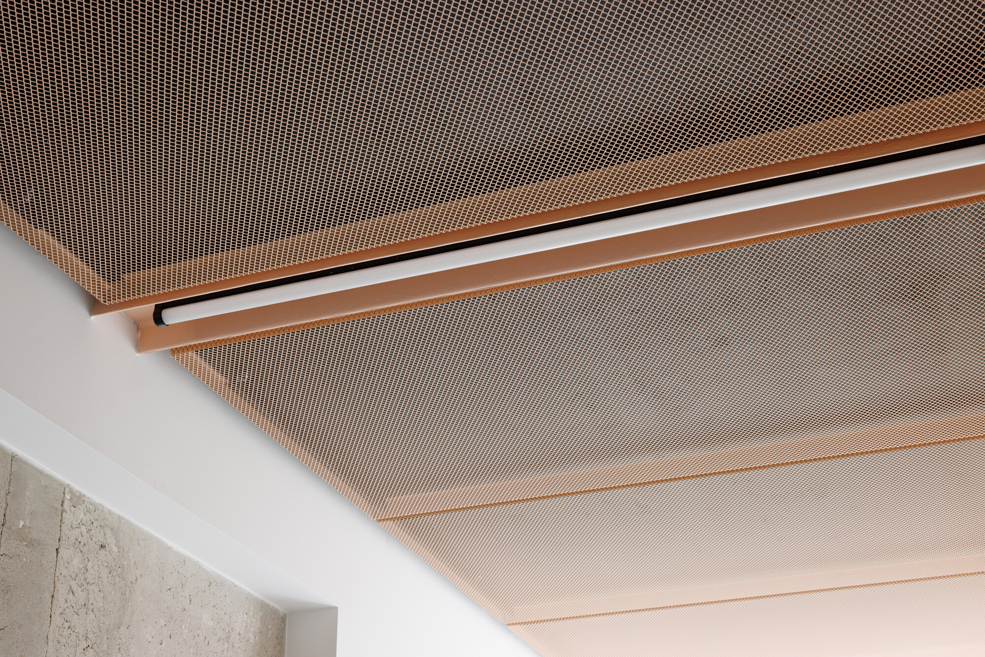 Close-up of a copper expanded mesh ceiling with a linear light fixture, meeting a white wall and raw concrete