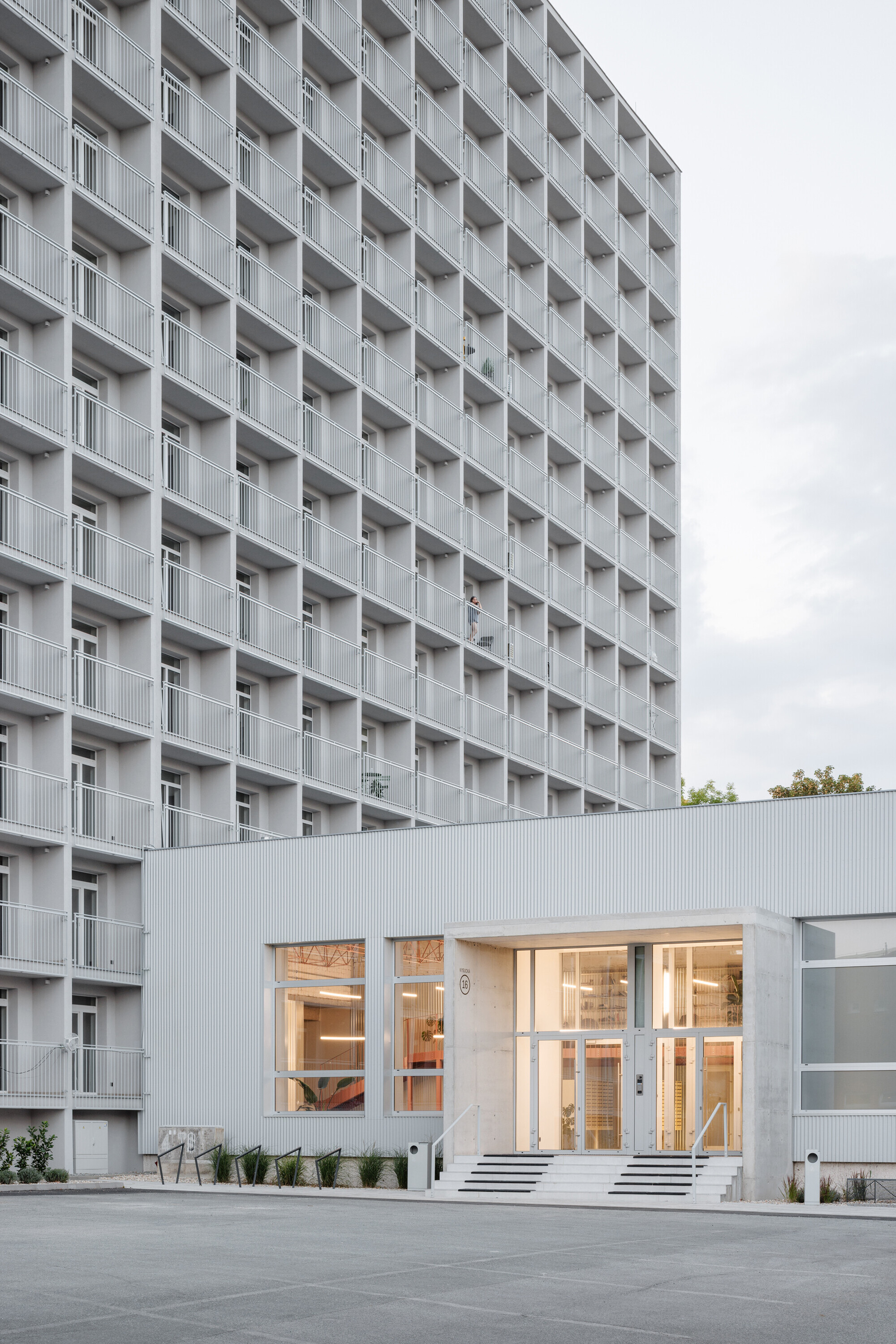 Gridded residential tower with deep balconies rising behind a low corrugated metal entrance block under an overcast sky