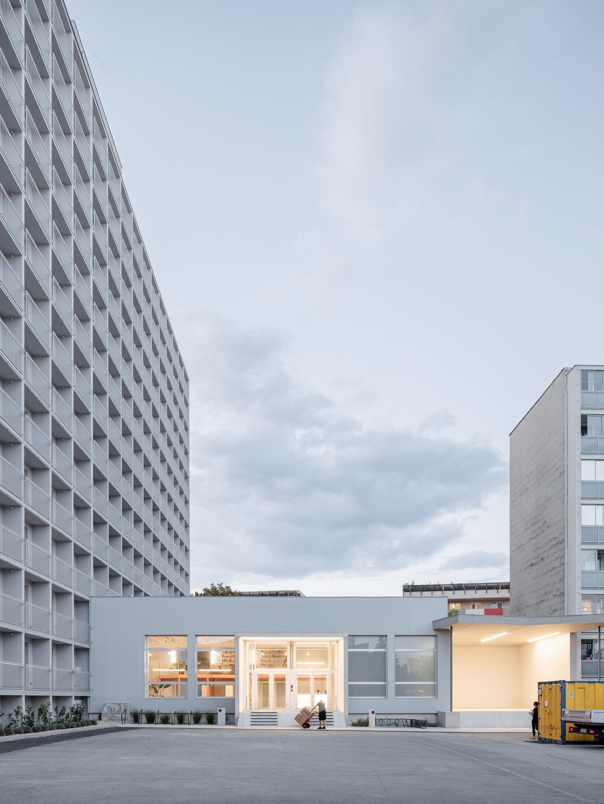 Low glazed entrance pavilion between a gridded slab block and a concrete tower, with a worker wheeling a trolley at dusk